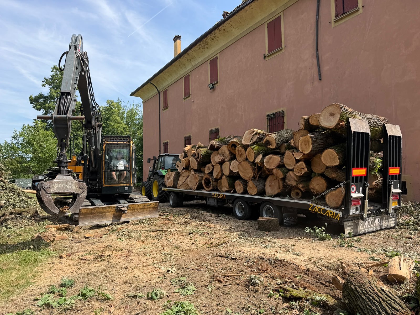 Escavatore e camion carico di tronchi di legno vicino a un edificio con pareti rosa e finestre con grate