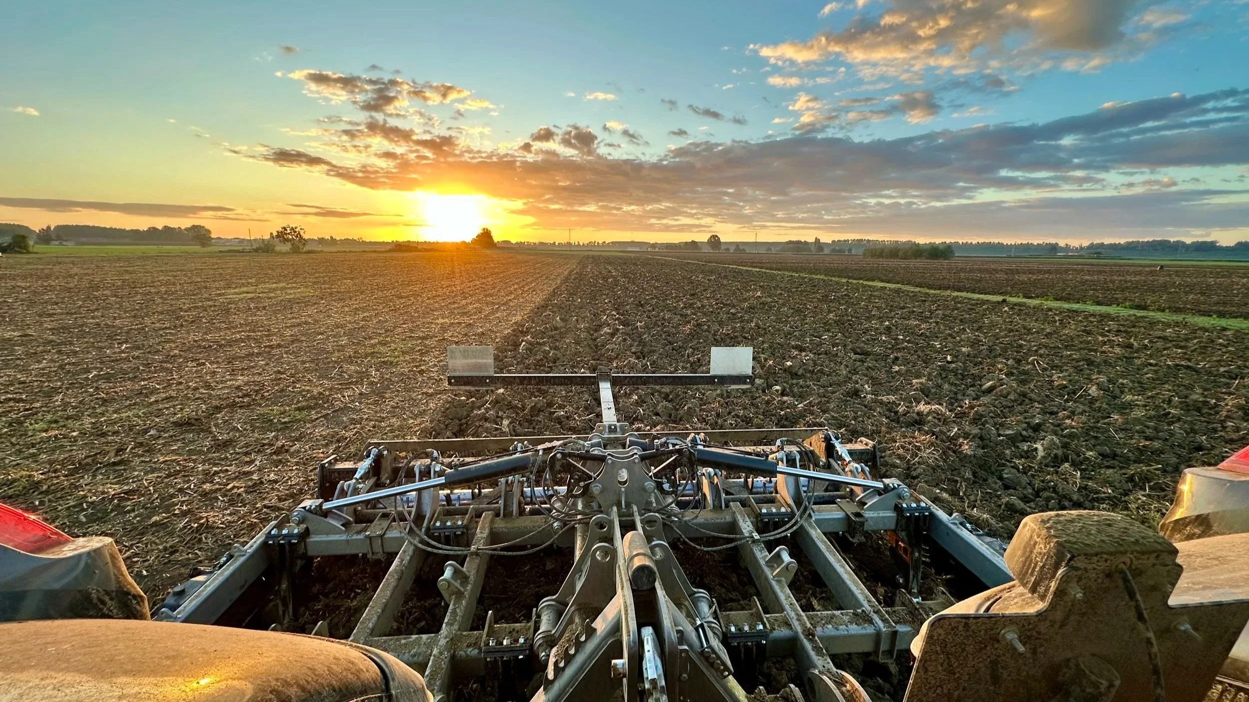 Campo agricolo con una macchina agricola che lavora la terra durante il tramonto, cielo con nuvole e sole in basso sull'orizzonte.