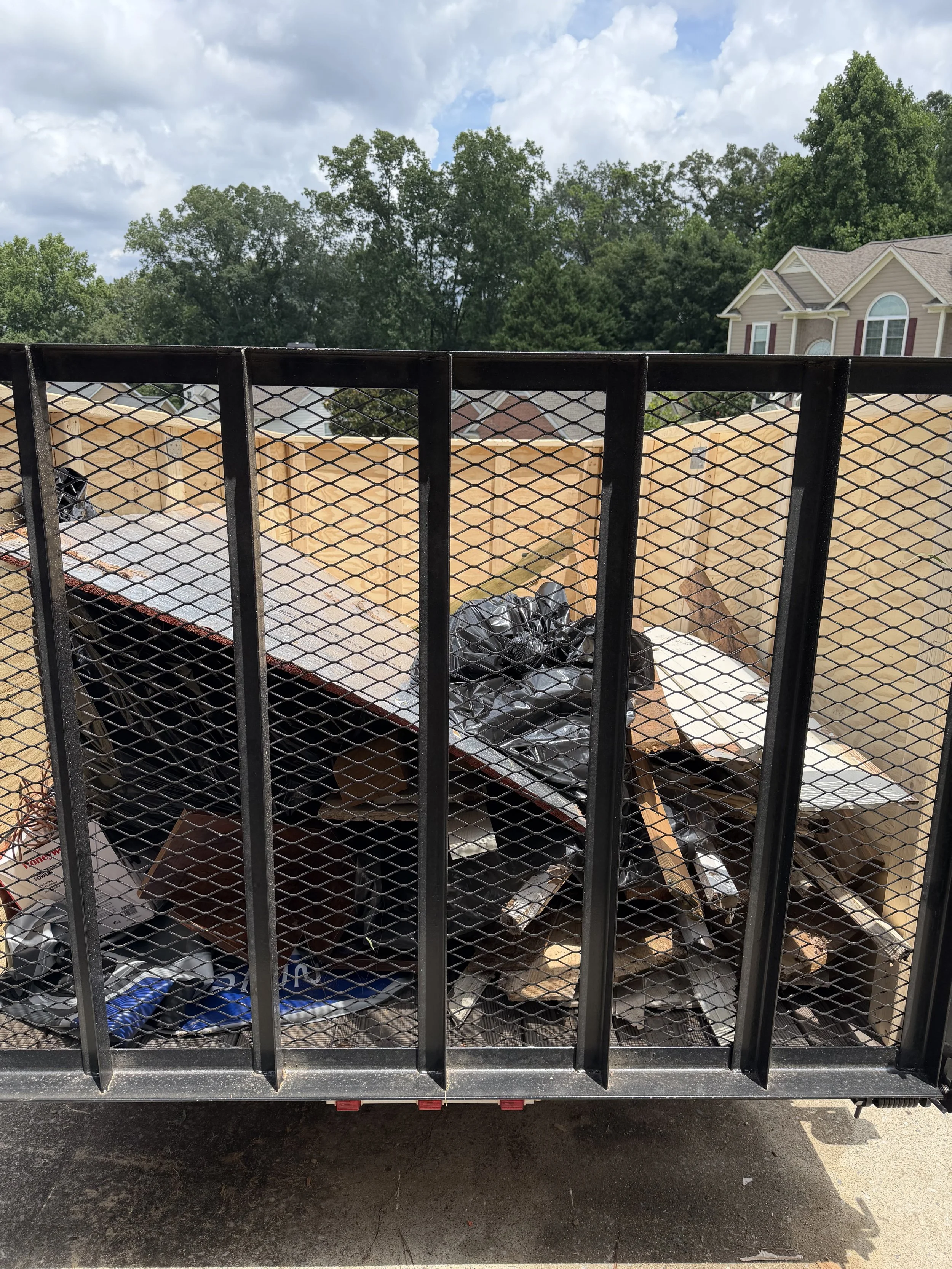 Back of a flatbed trailer filled with construction debris, including black plastic bags, wood, and cardboard, parked in an outdoor area with residential houses and trees in the background.