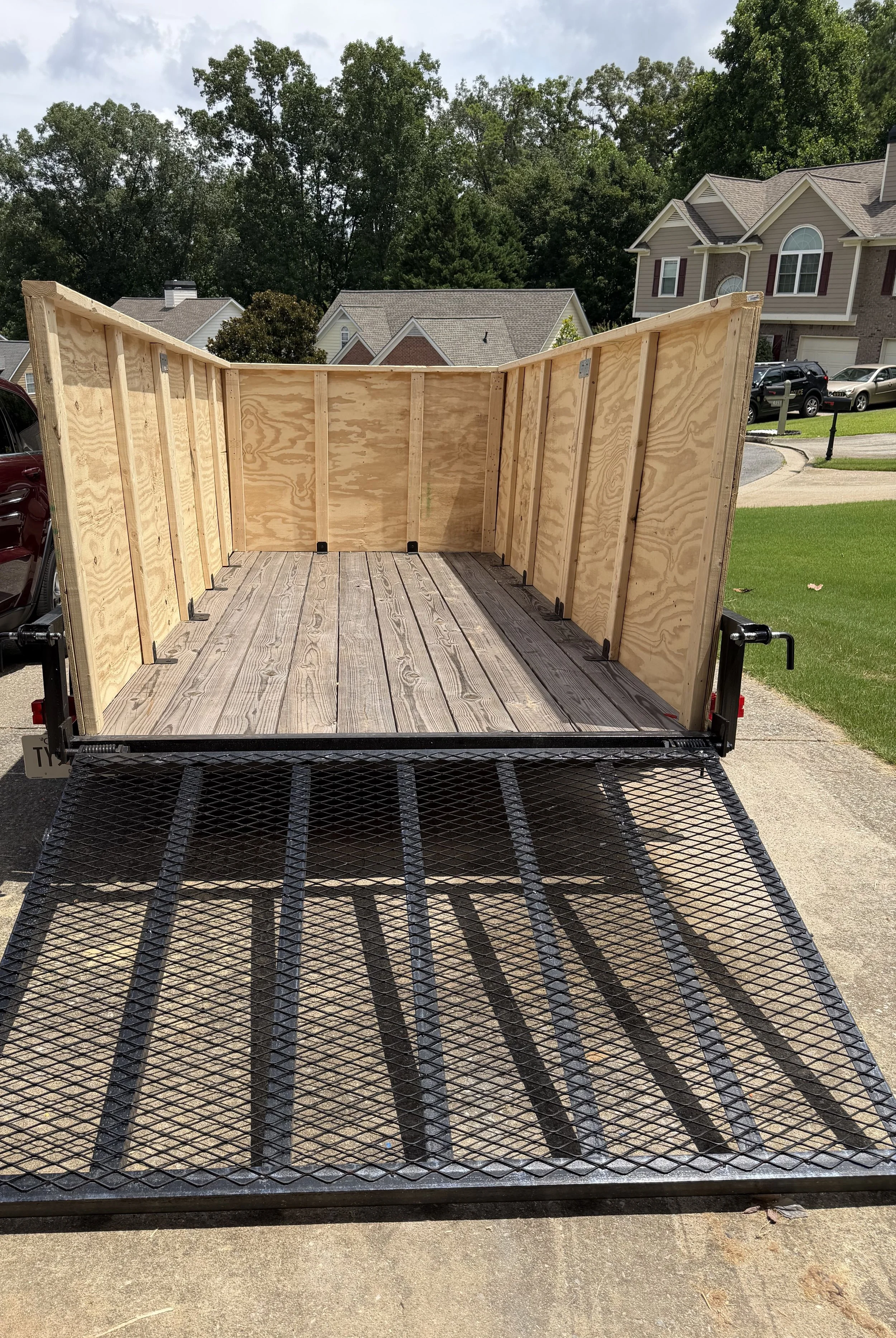A flatbed trailer with wooden sides parked on a driveway in a residential neighborhood. The trailer has a wooden floor and sides made of vertical plywood panels framed with wood. In the background, there are houses, trees, and parked cars.