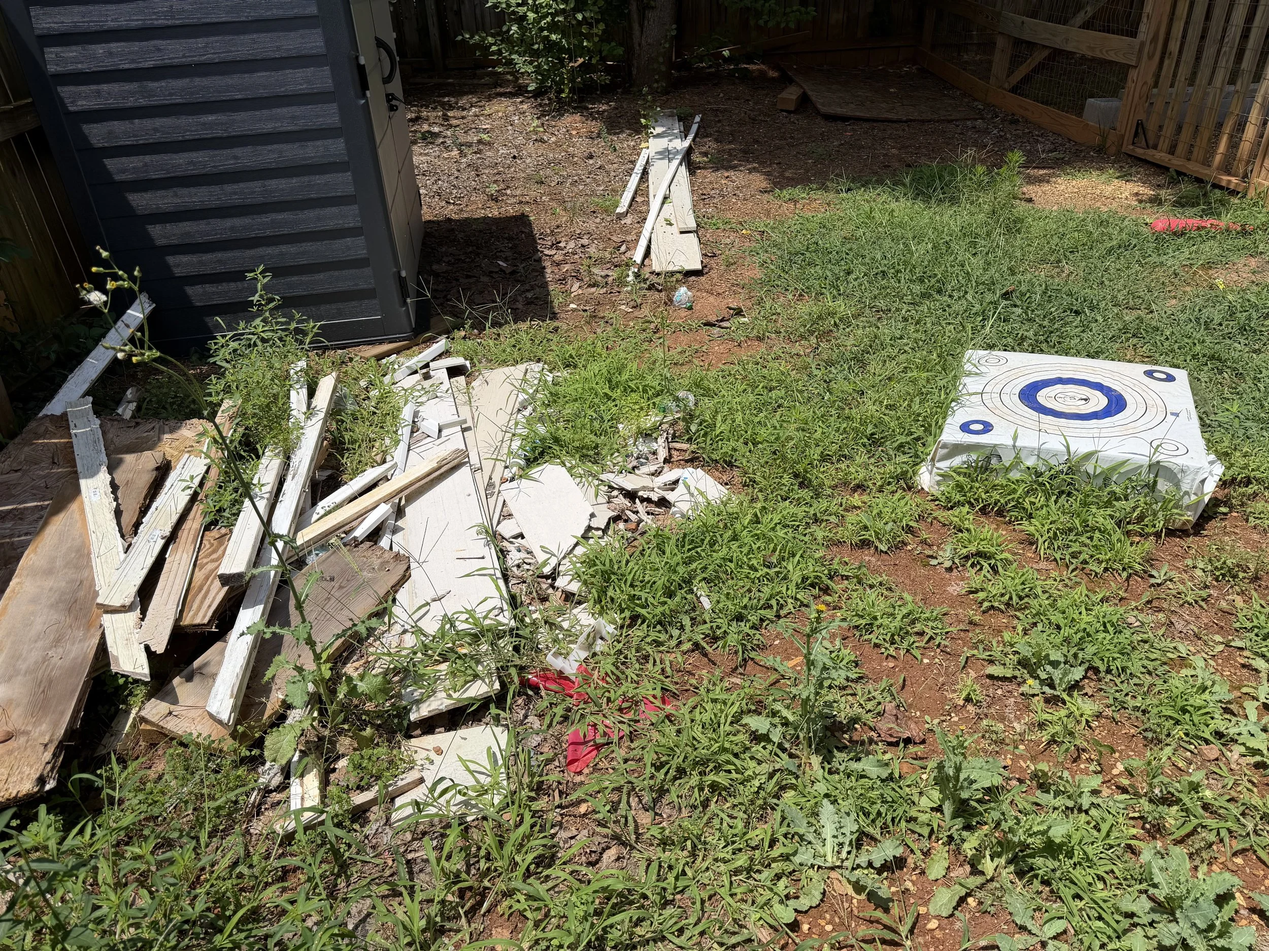 Backyard with broken wooden fence pieces, some greenery, and an archery target on the ground.
