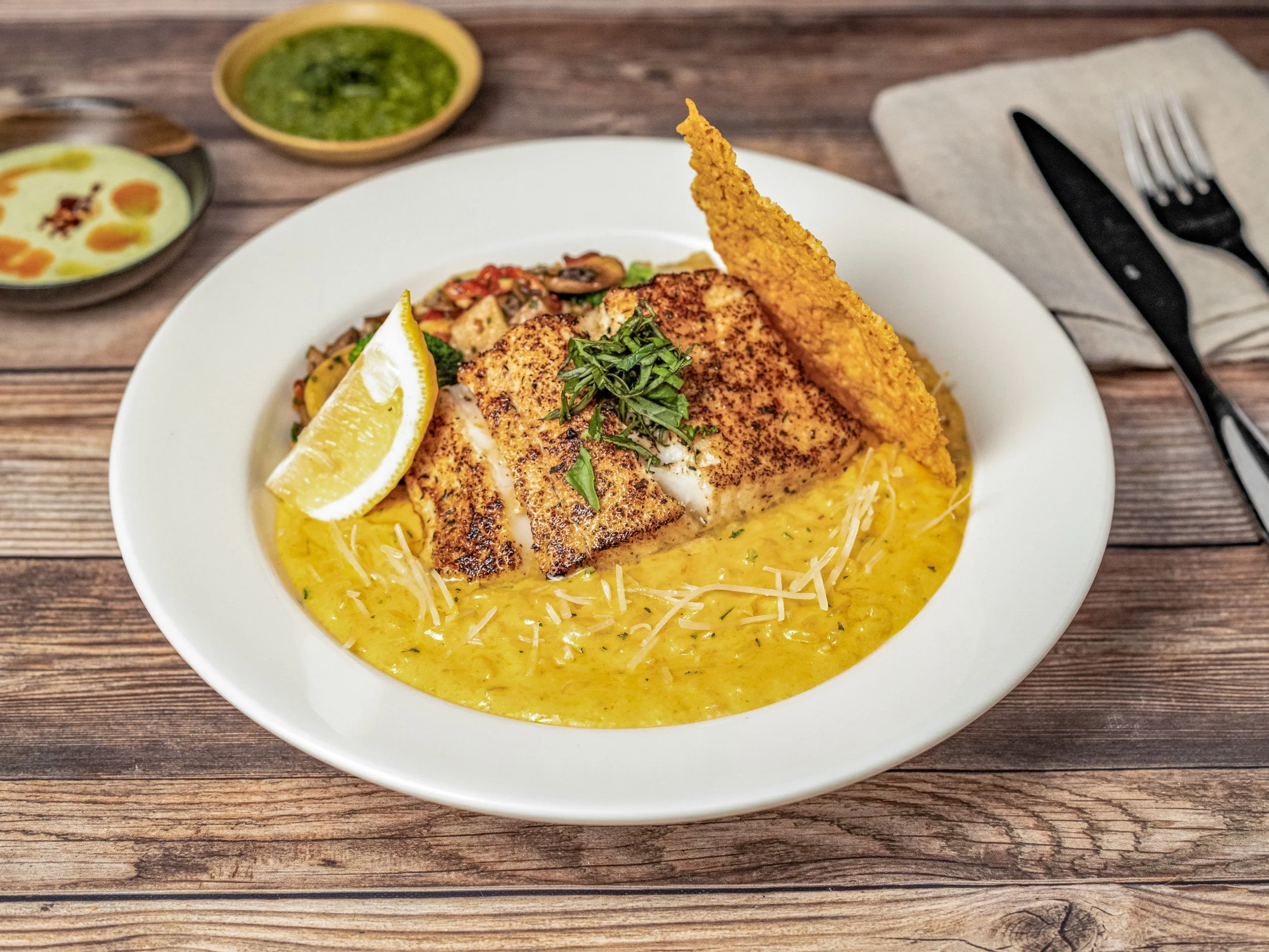 A white plate of grilled fish fillet topped with chopped herbs, served over yellow rice, with a lemon wedge on the side, accompanied by two sauces and placed on a wooden table.