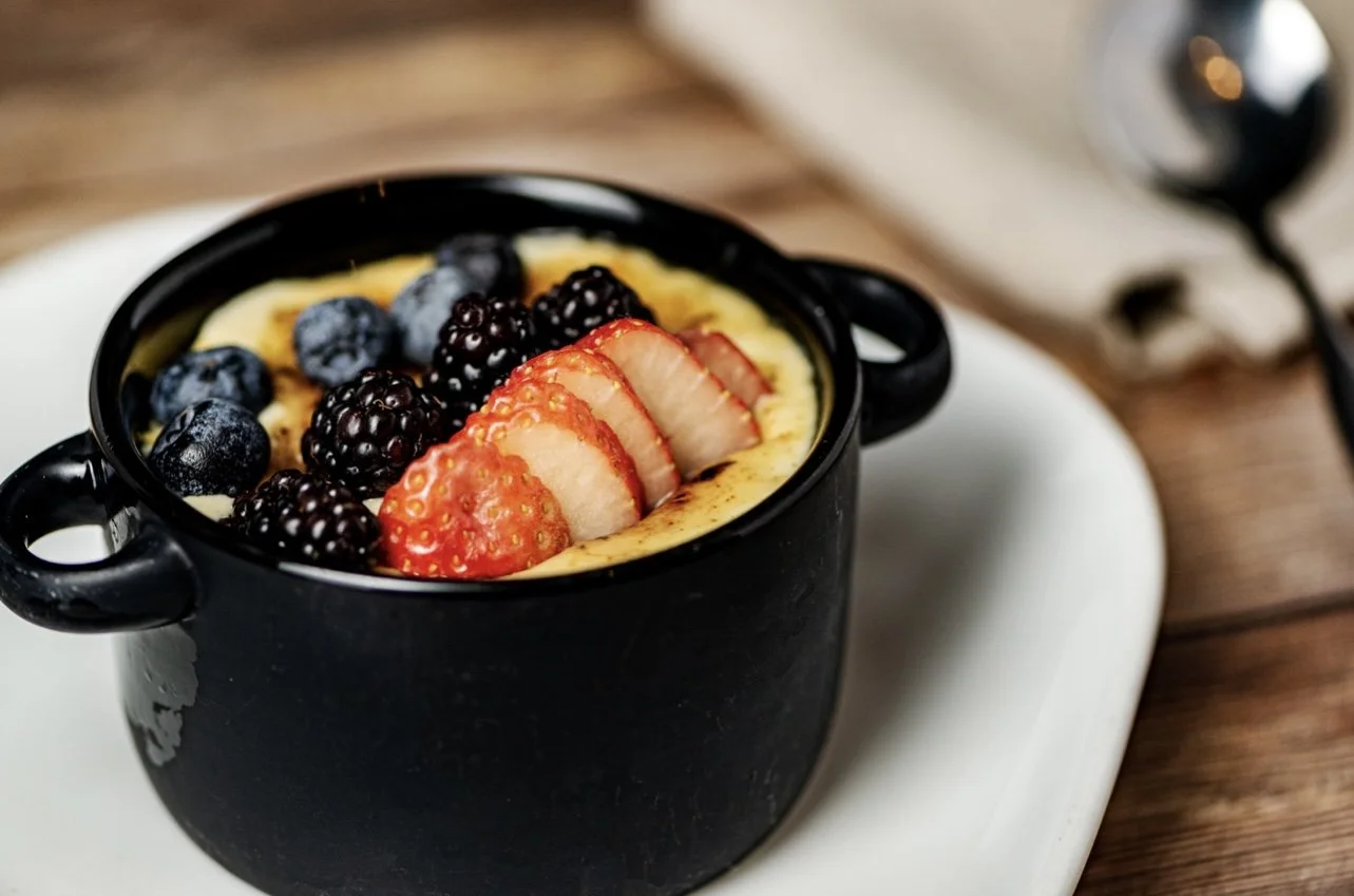 Black mug filled with assorted fresh berries and sliced strawberries on top of a custard or pudding dessert, placed on a white plate on a wooden surface.
