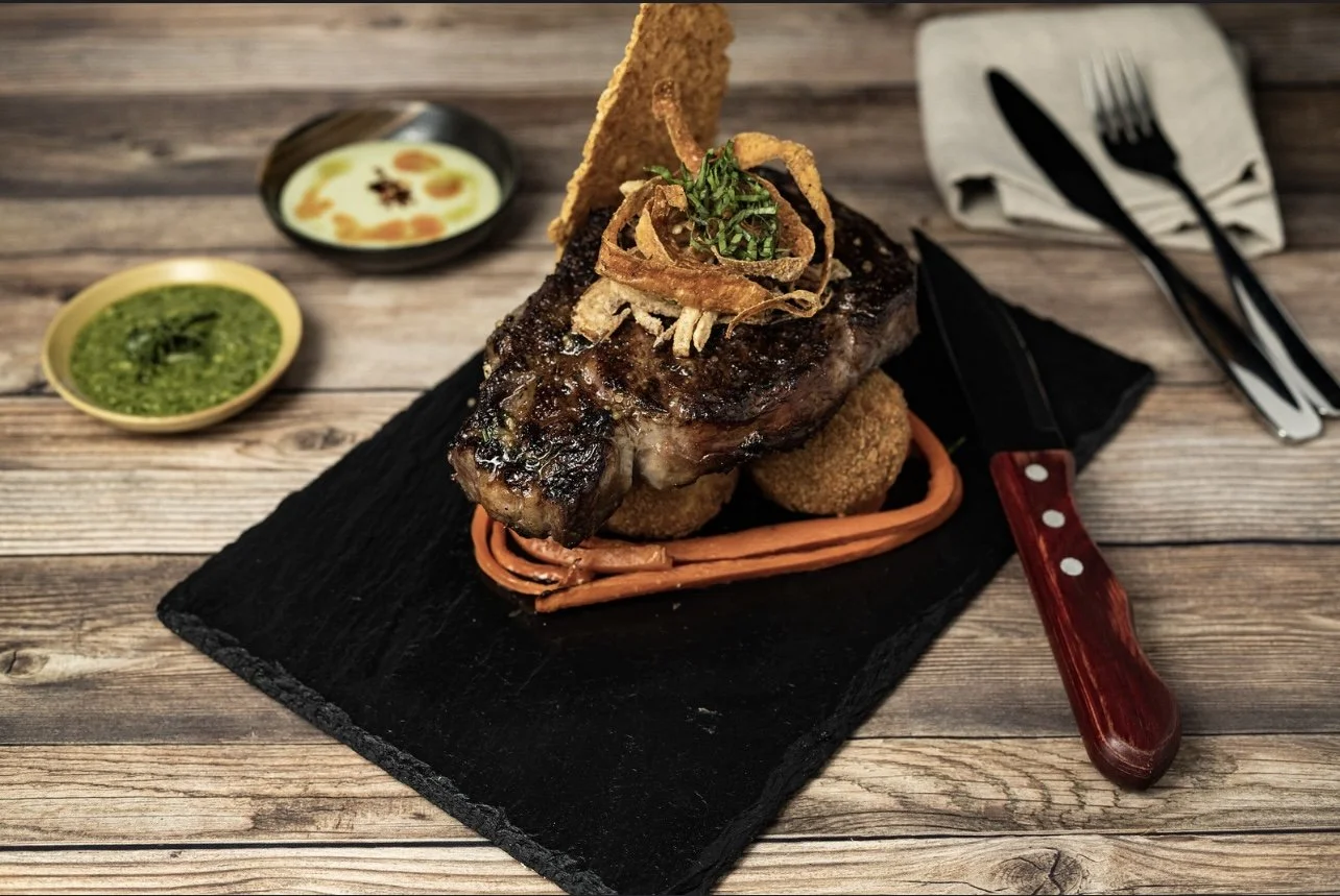 Grilled steak topped with crispy onion strings, garnished with herbs, served on a bed of fried croquettes and carrot puree, with dipping sauces on side, placed on a black slate plate.