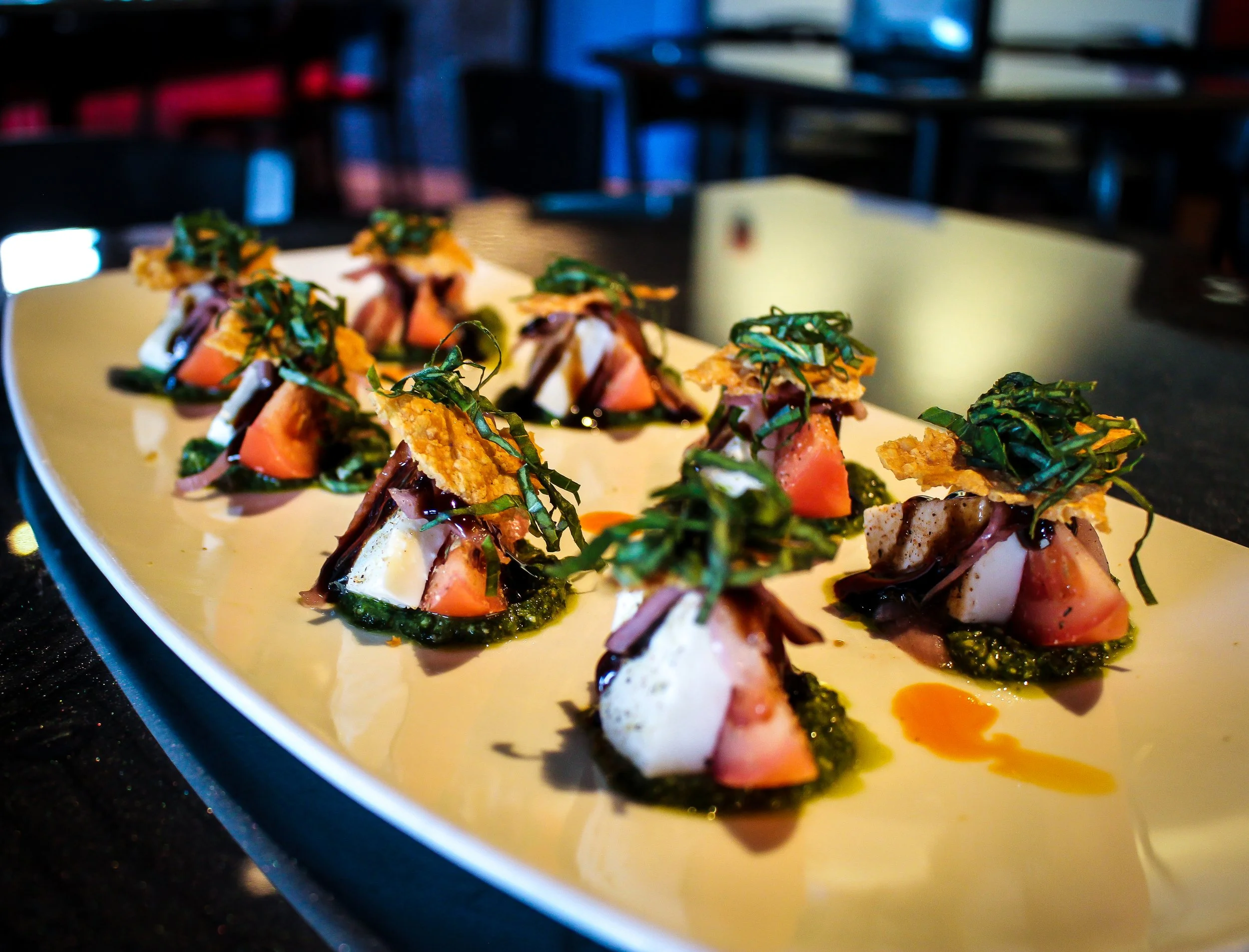 Close-up of multiple gourmet appetizers on a white plate, garnished with herbs and drizzled with sauce, on a dark countertop in a restaurant setting.