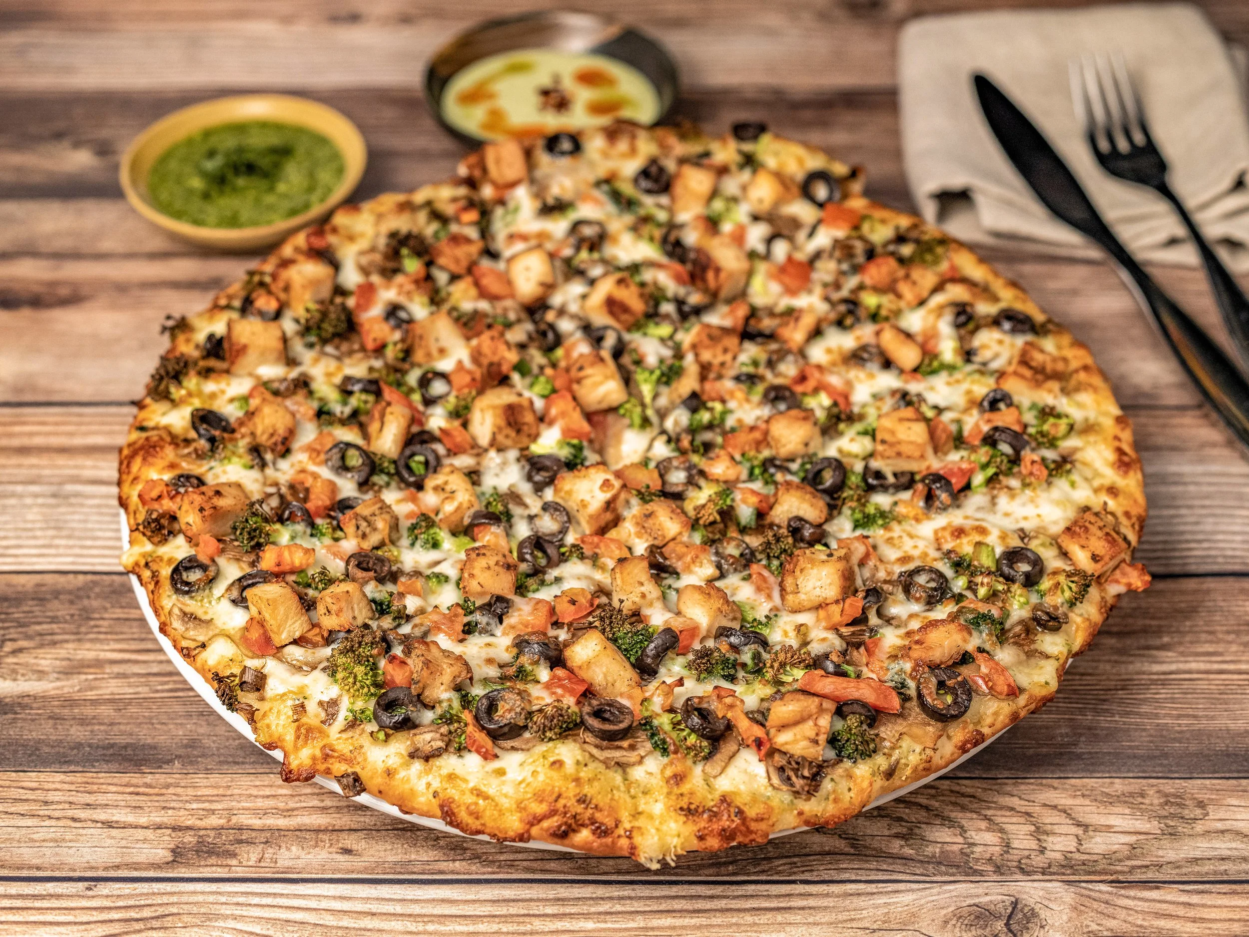 A large pizza topped with chicken pieces, black olives, broccoli, and other vegetables on a wooden surface with two small bowls of dipping sauces and black cutlery.