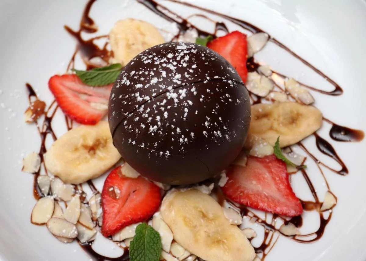 Chocolate mousse dessert with strawberries, banana slices, chopped nuts, mint leaves, and chocolate drizzle on a white plate.
