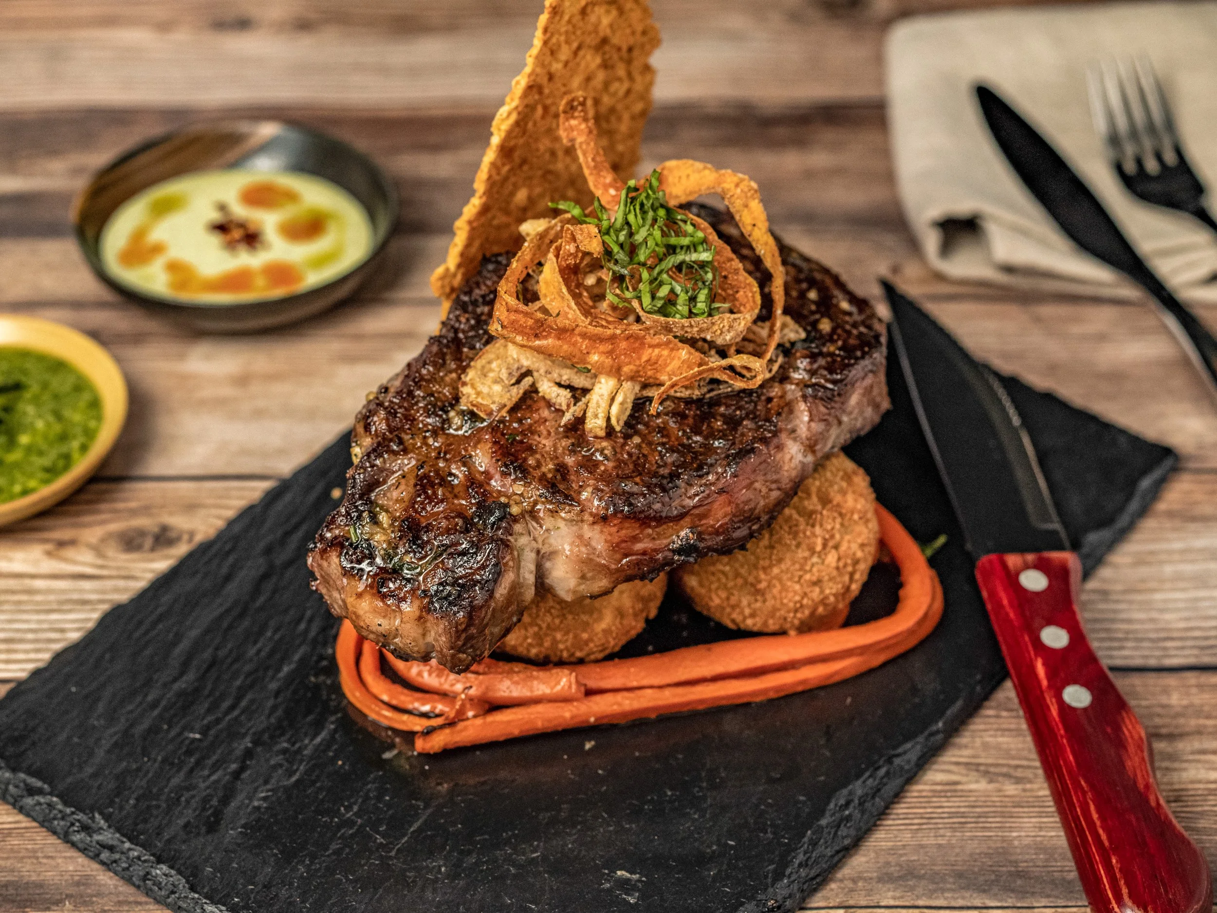 A large beef steak topped with crispy onion strings and green herbs, served on a black slate platter with side sauces in small bowls.