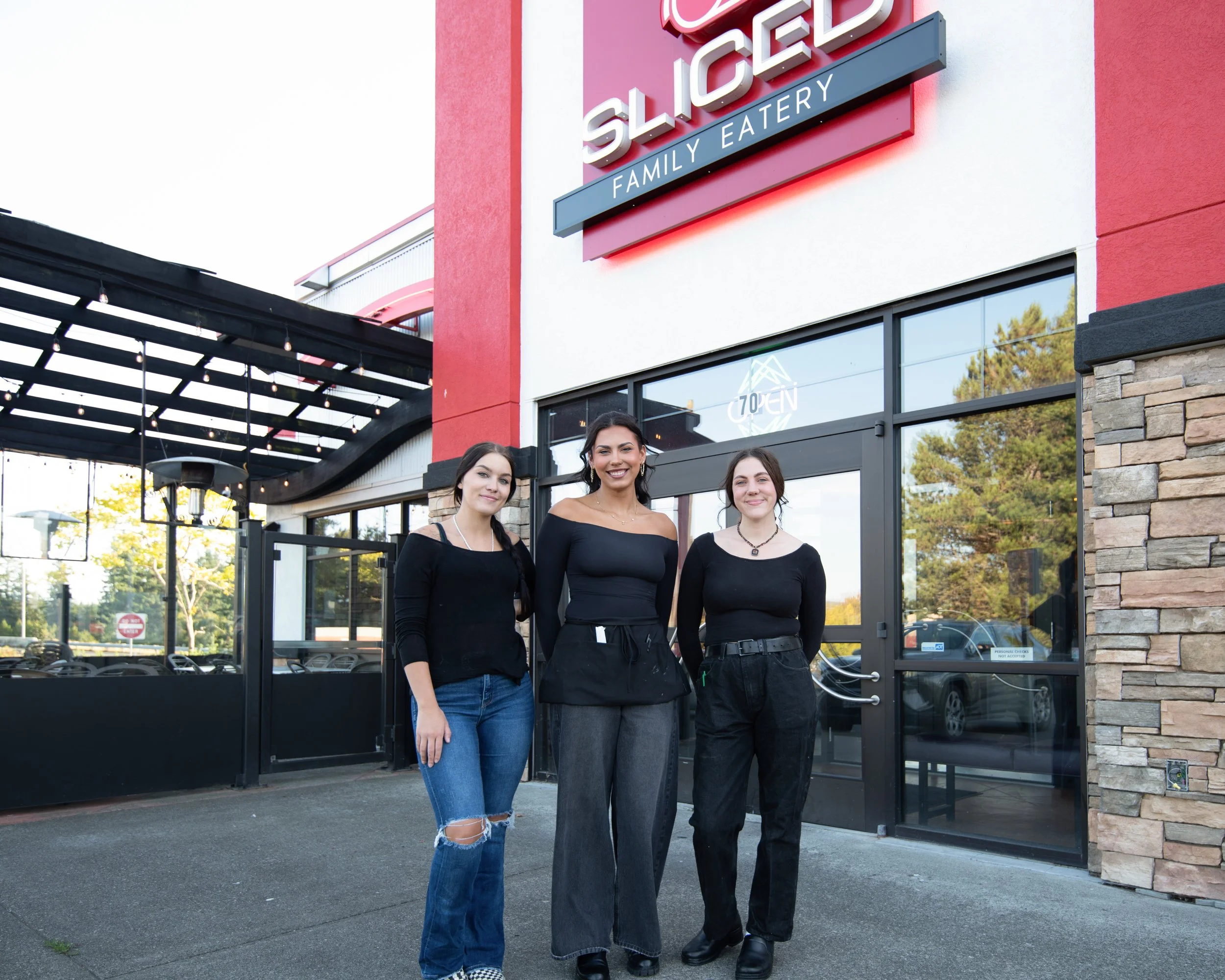 Four women standing outside a family eatery restaurant with red and white exterior, glass doors, and a sign that says 'SLICE'D FAMILY EATERY.' The women are smiling and dressed in black tops and jeans.