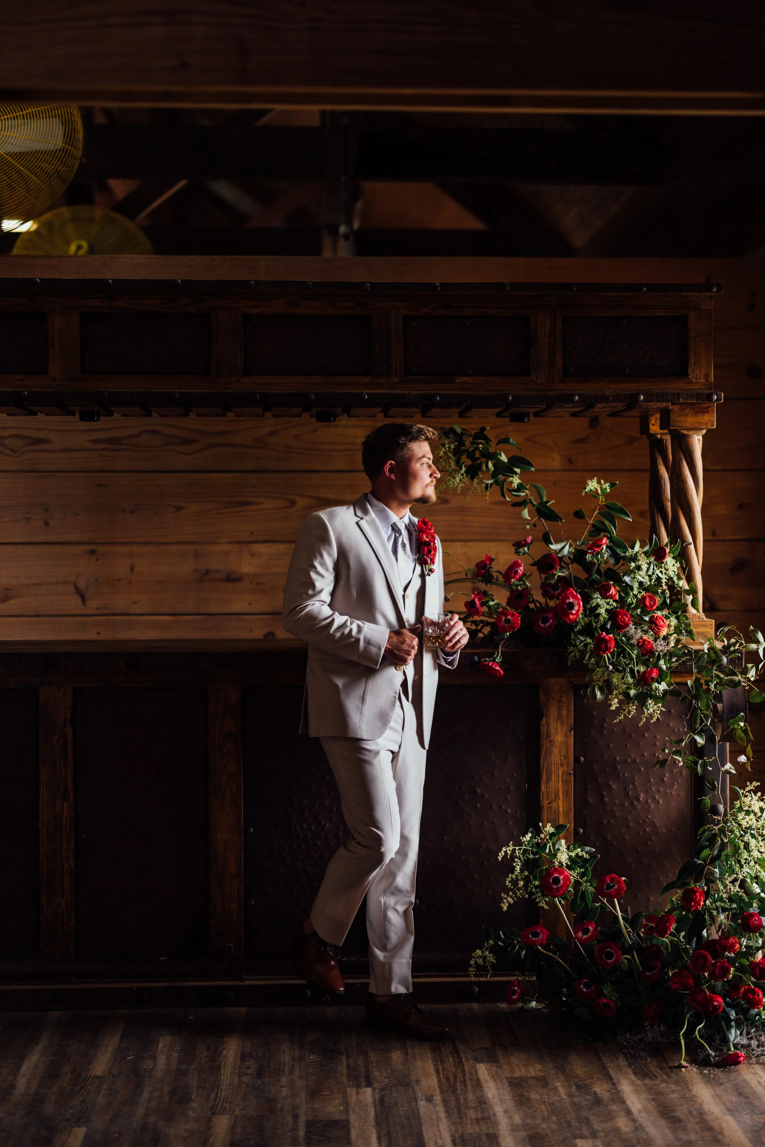 A groom in a white suit with a red flower boutonniere holding a drink, standing next to a floral arrangement with red and white flowers, in a wooden interior setting with warm lighting.