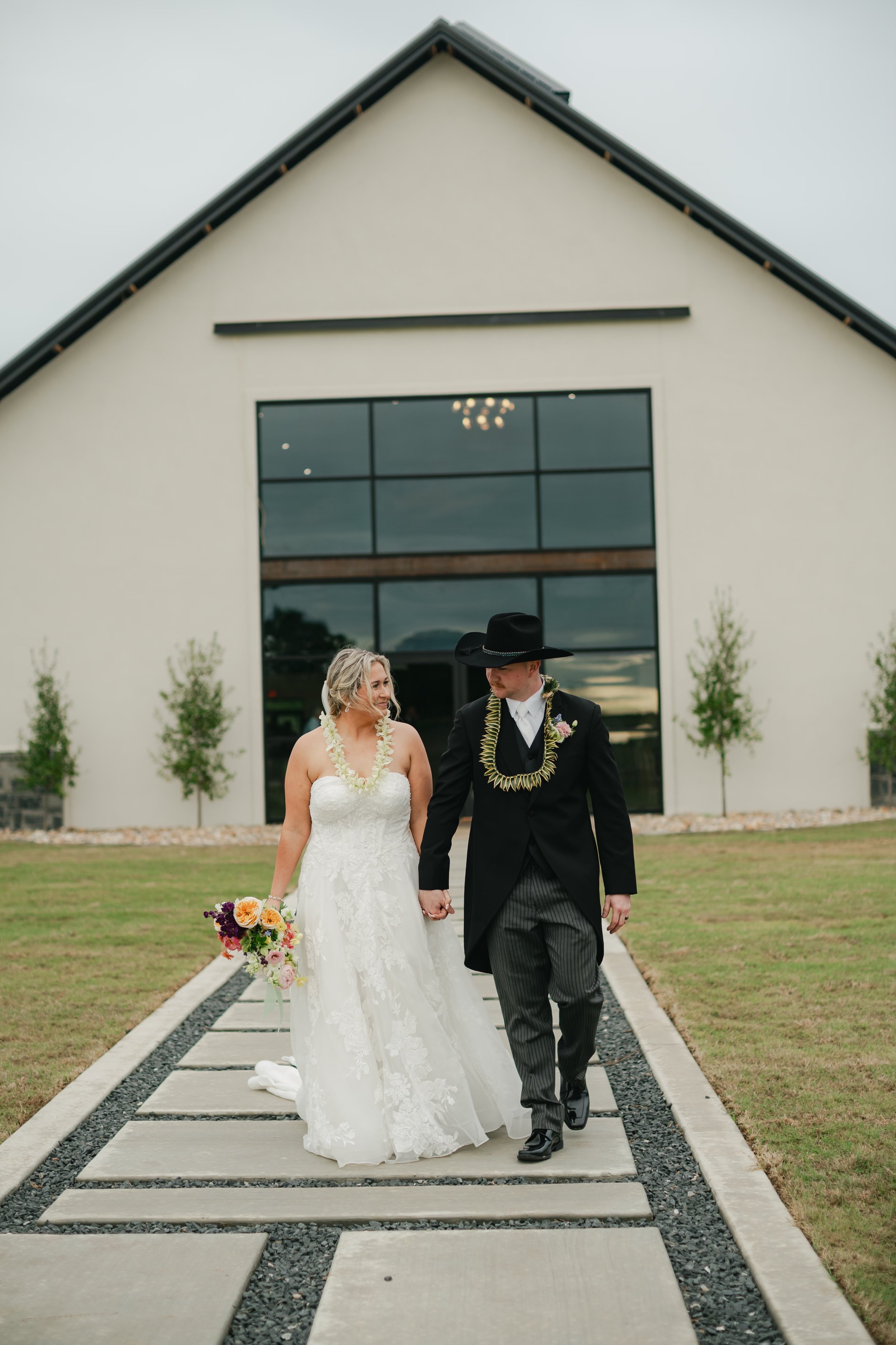 A bride and groom walking hand in hand on a walkway outside a modern building, holding a bouquet of flowers, dressed in wedding attire. The bride wears a white train and the groom is in a black suit with a lei around his neck.
