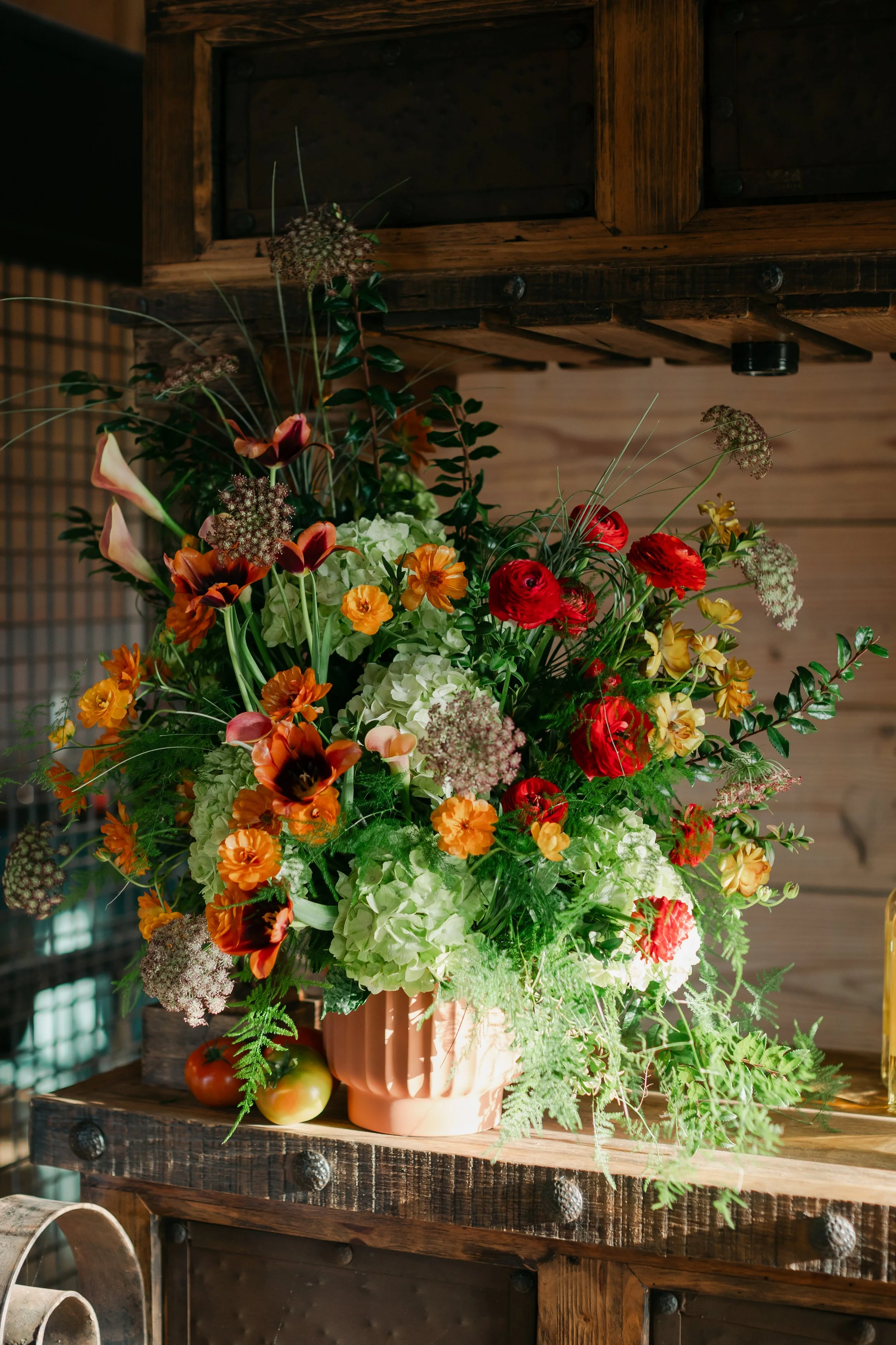 A large floral arrangement in a pink vase on a wooden table, with assorted flowers and greenery including calla lilies, ranunculus, and hydrangeas, set against a wooden wall and ceiling.