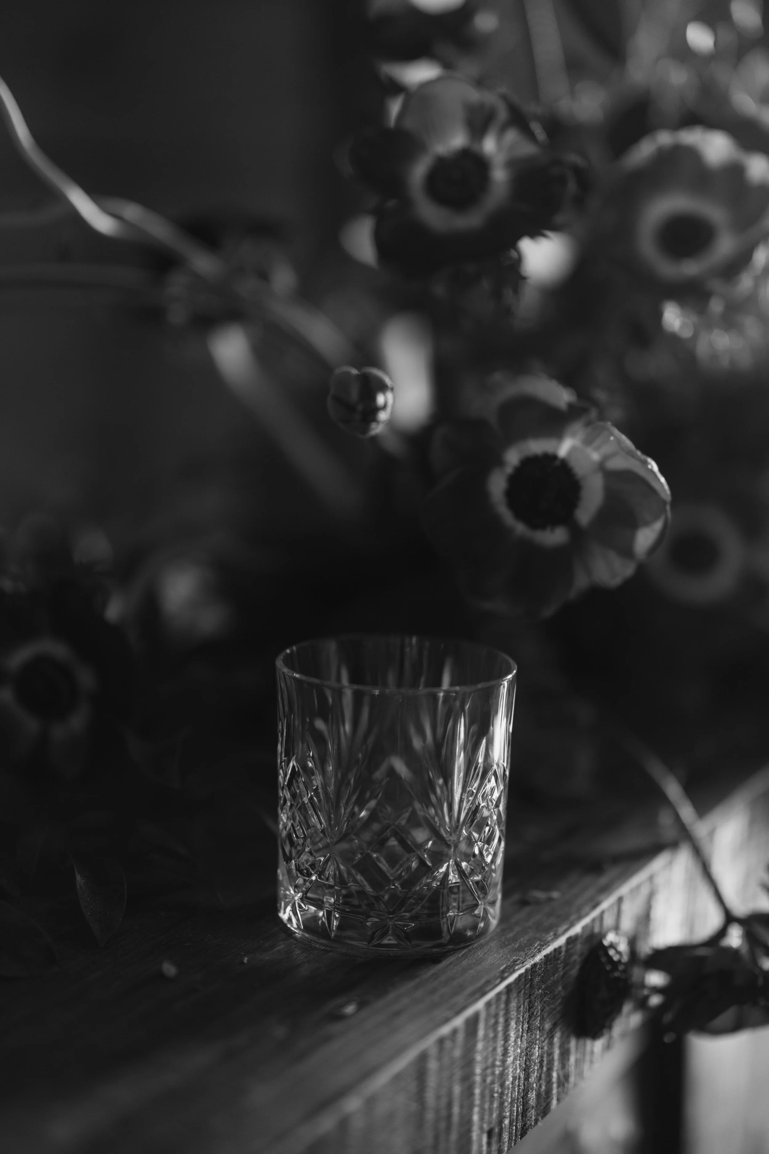 A clear, faceted glass cup is on a wooden surface in a black and white photo. Behind the cup, there are blurred flowers or plant-like objects on a vertical surface.