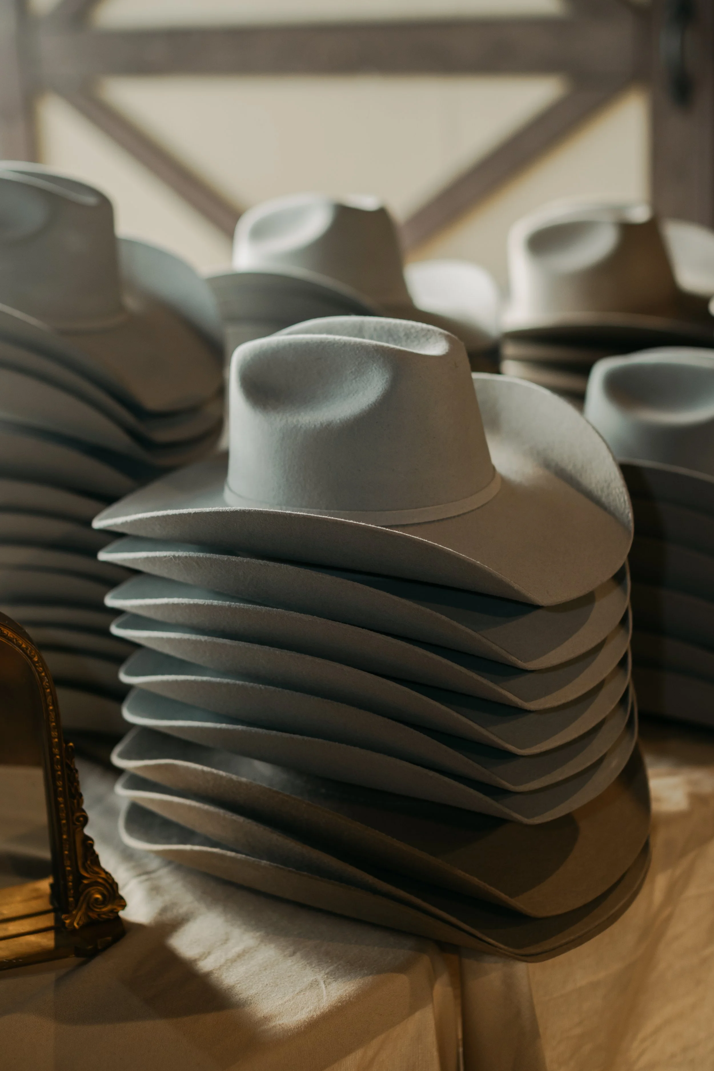 A stack of white cowboy hats arranged in a pattern with a tilt, with a decorative mirror and a table with a cloth beneath it in the background. Cavendrer's Hat Bar for Wedding favors