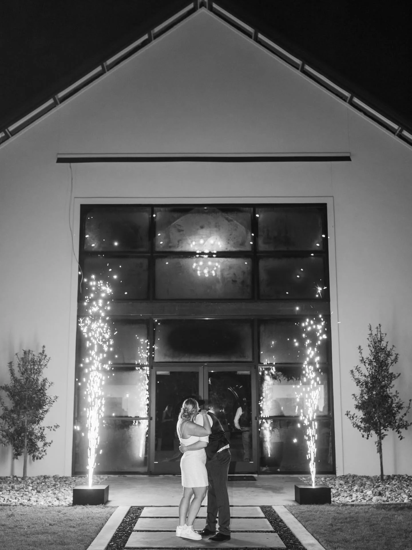 A couple dancing in front of a modern building with large glass windows, illuminated by COLD SPARK sparklers, at night.