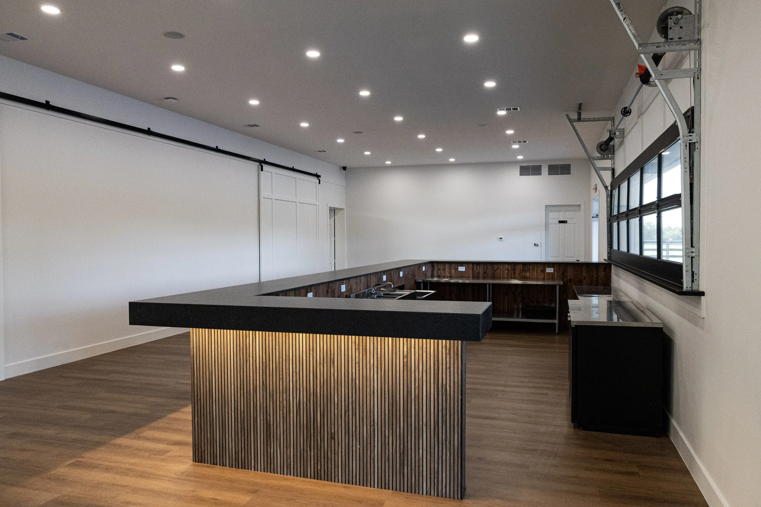 Empty modern kitchen with a dark gray island, white walls, wood flooring, and ceiling lights. The right wall has large windows and a garage door.