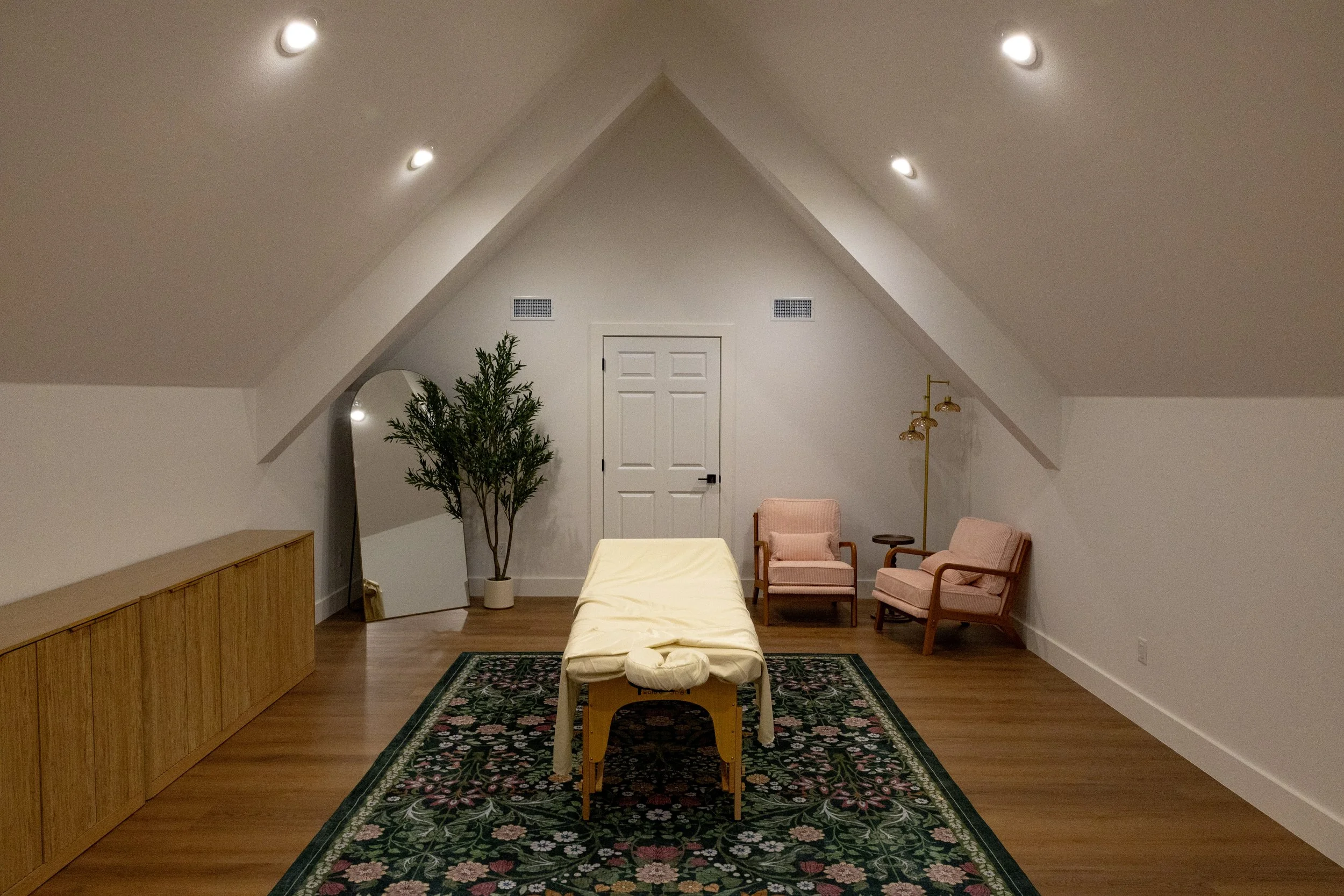 A cozy room with a sloped ceiling illuminated by four recessed lights. The room features a white door, a potted plant, a large mirror, two pink armchairs, a wooden sideboard, a gold floor lamp, and a floral patterned rug.