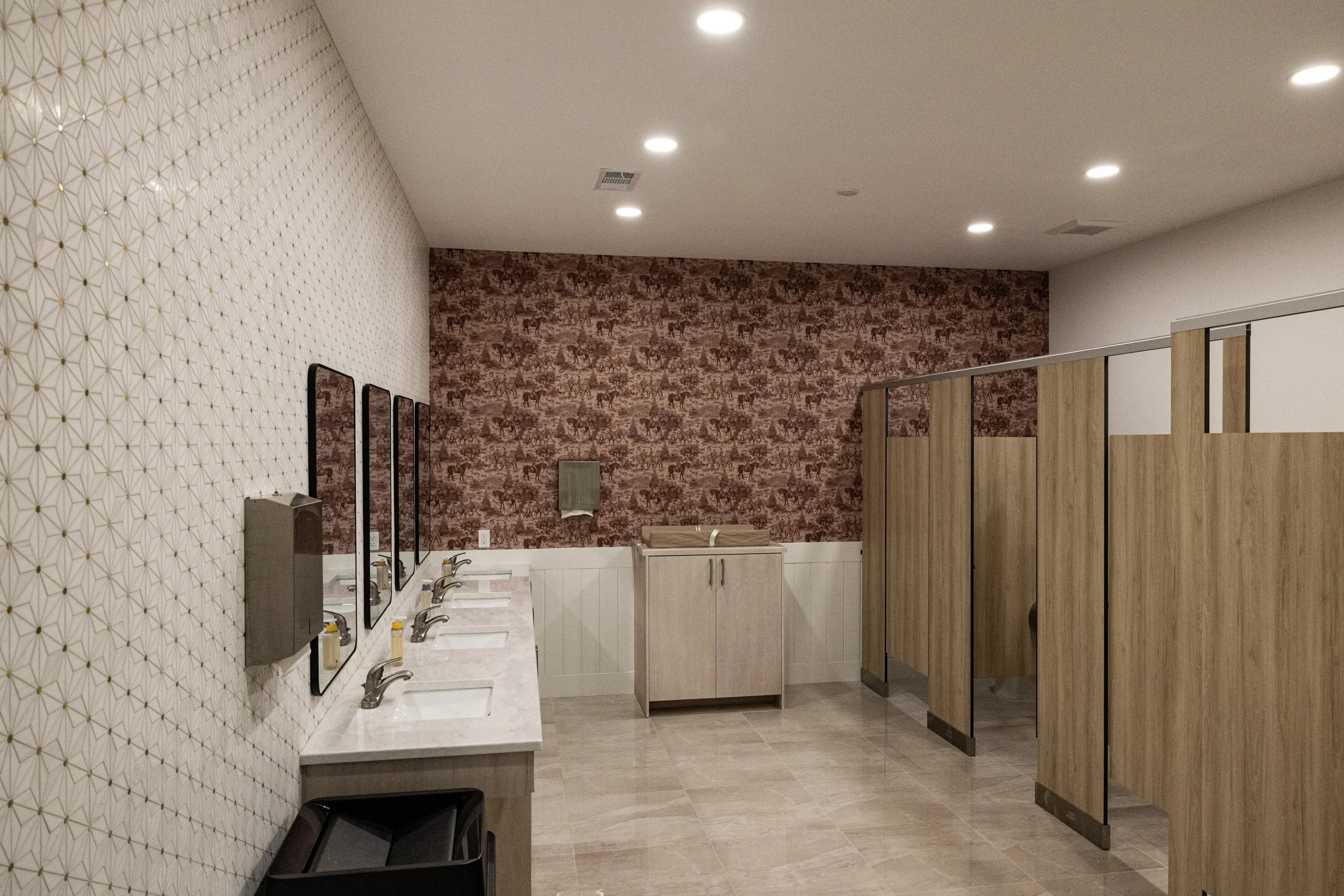 Public restroom with multiple sinks, mirrors, soap dispensers, and wooden partitioned stalls. Wallpaper with wild animal print on the back wall, and geometric patterned wallpaper on the side wall.