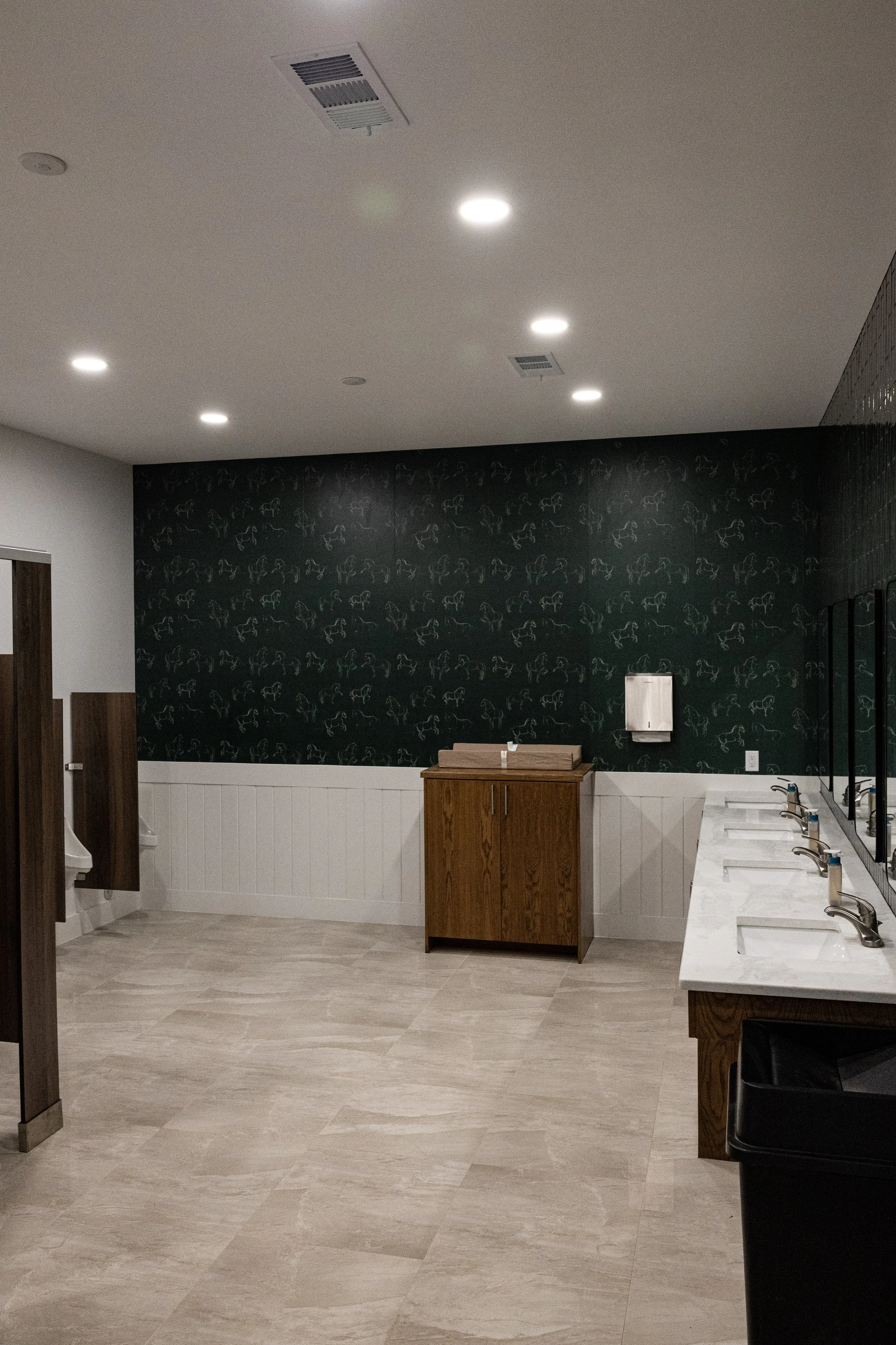 A modern public restroom with beige wood-look flooring, a white wainscot and a black wallpapered accent wall featuring a simple line drawing of a horse. The white marble countertop with sinks is visible on the right side. A small wooden cabinet is in