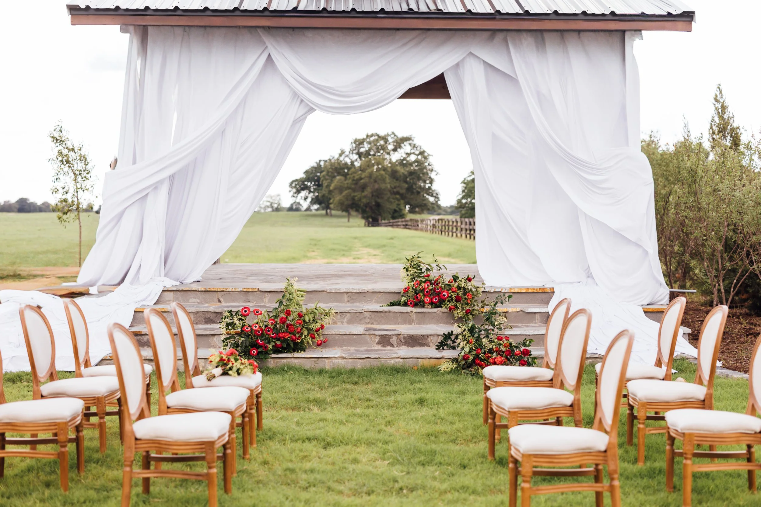 An outdoor wedding ceremony setup with rows of white chairs with wooden frames facing a decorated arch with white drapes and red flowers at the base, in a grassy field with trees in the background.