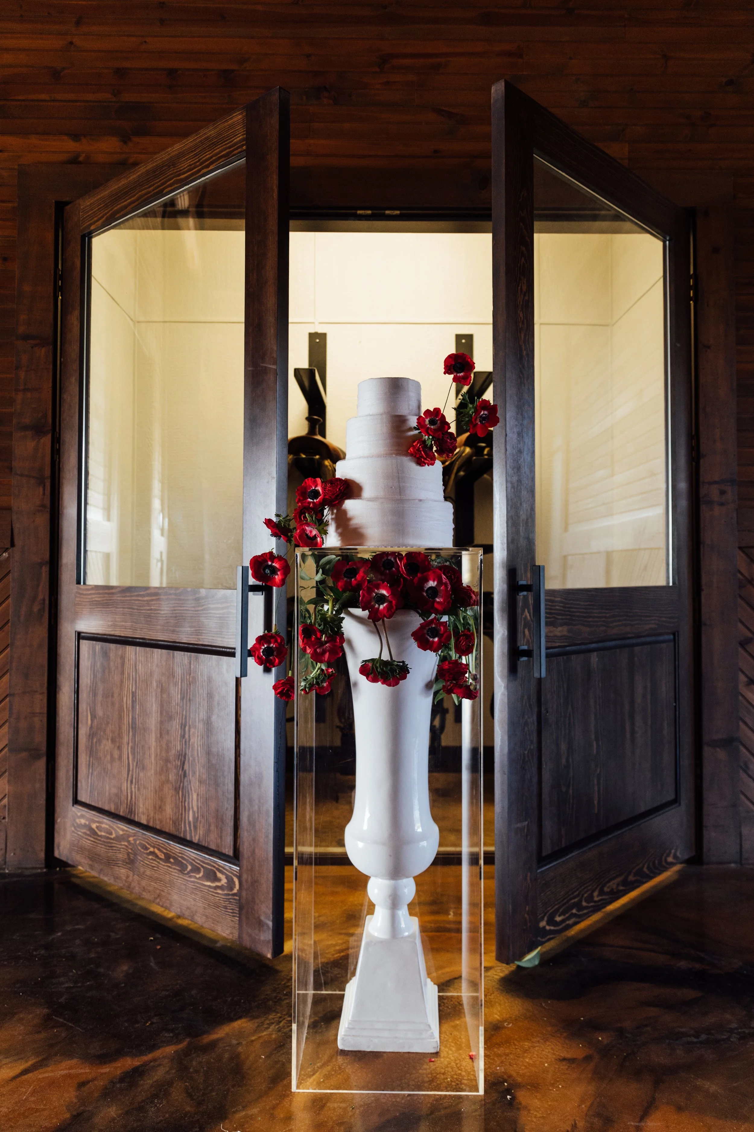 A white pedestal vase filled with red flowers is displayed inside a glass case in front of a wooden door, with a wedding cake visible in the background.