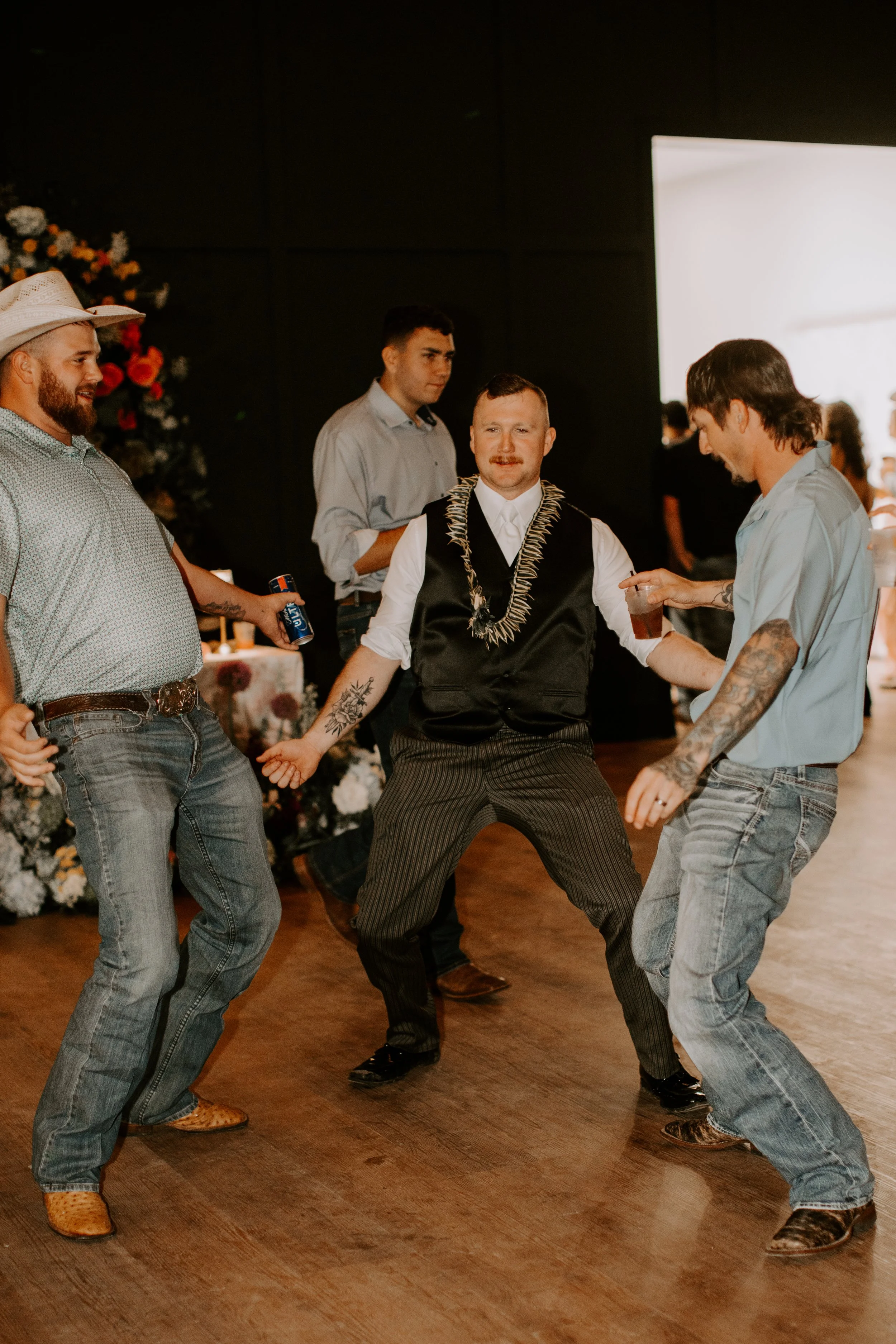 Group of five men dancing together indoors, with one in the center wearing a lei and striped pants, and others wearing casual clothes, with a floral backdrop and bright window in the background.