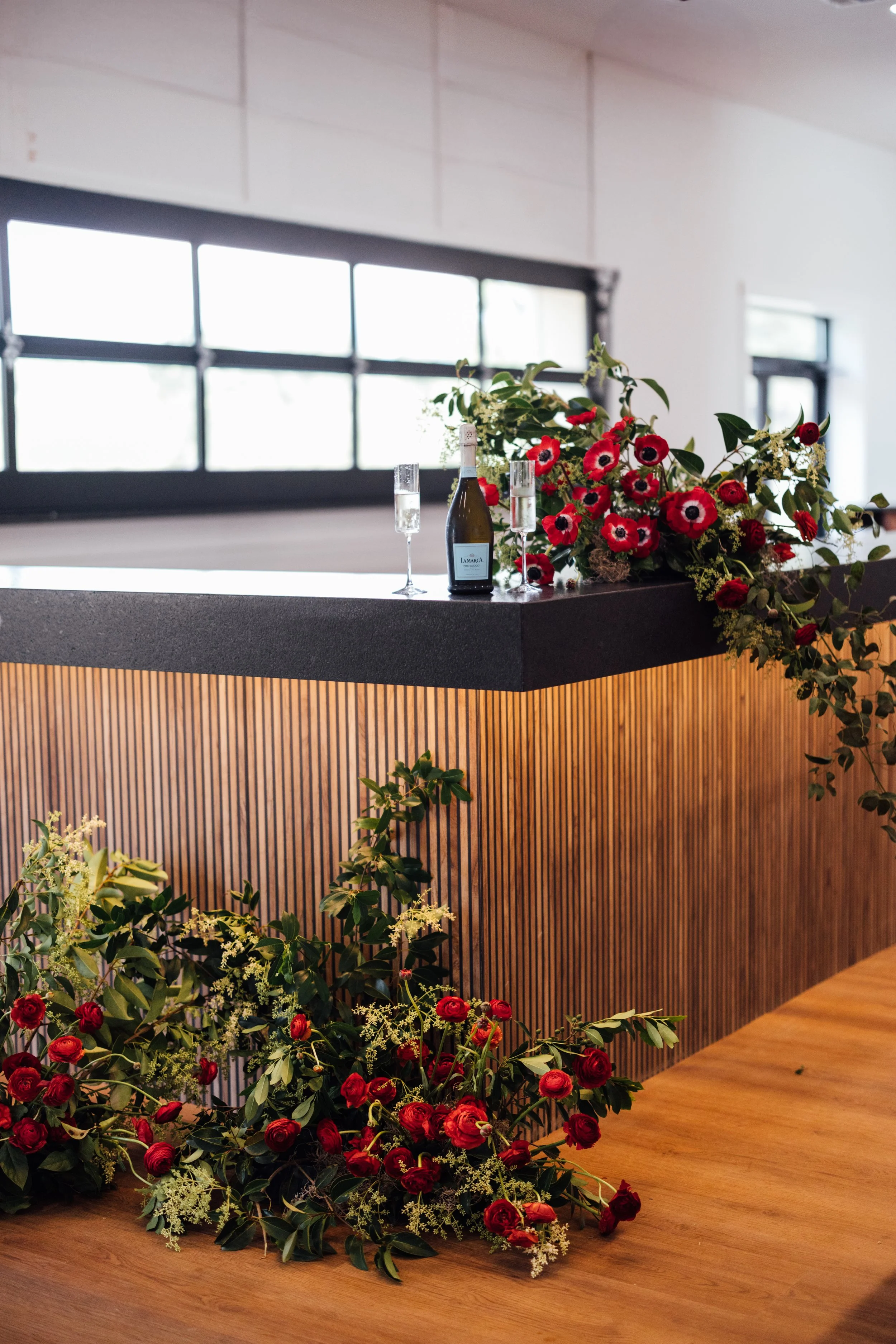 Custom Large Bar top for weddings and events with a bouquet of red and white flowers, a bottle of wine, and two glasses. Large windows in the background provide natural light.