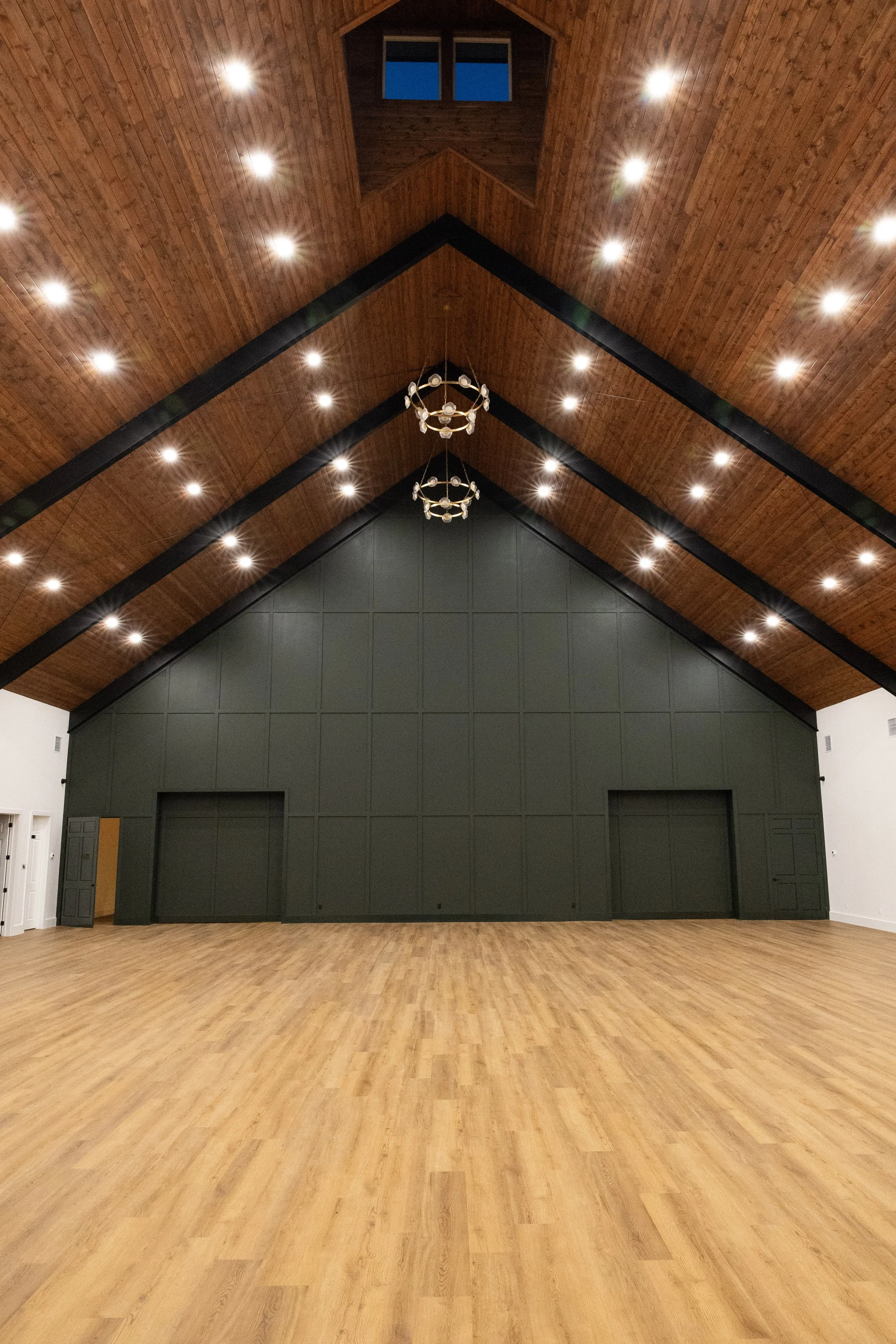 Interior view of a large wedding venue with a high wooden ceiling, ceiling lights, chandeliers, a dark green wall with paneling, and wooden flooring.