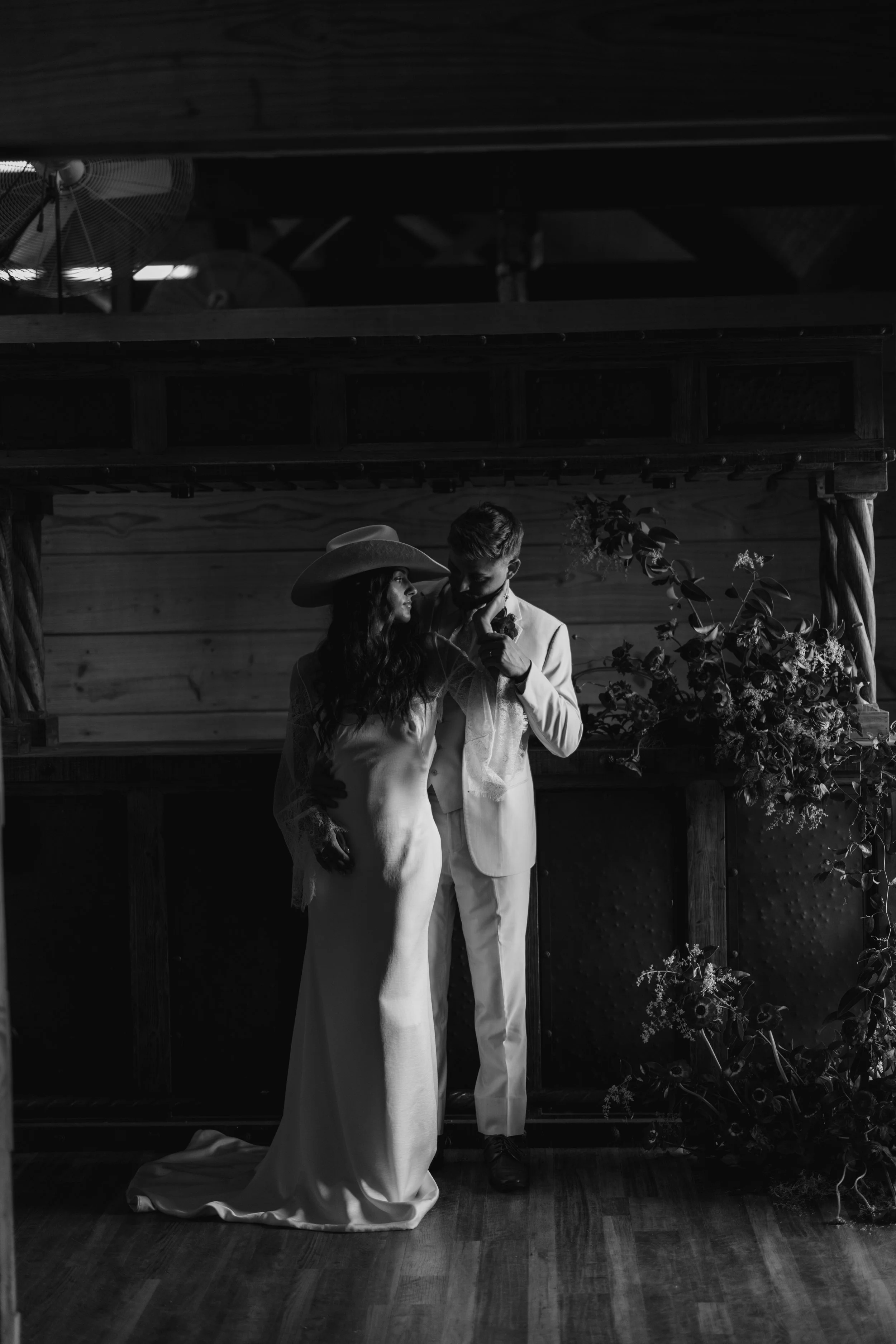 A bride and groom are dancing together at a rustic wedding venue, with a wooden backdrop and floral decorations.