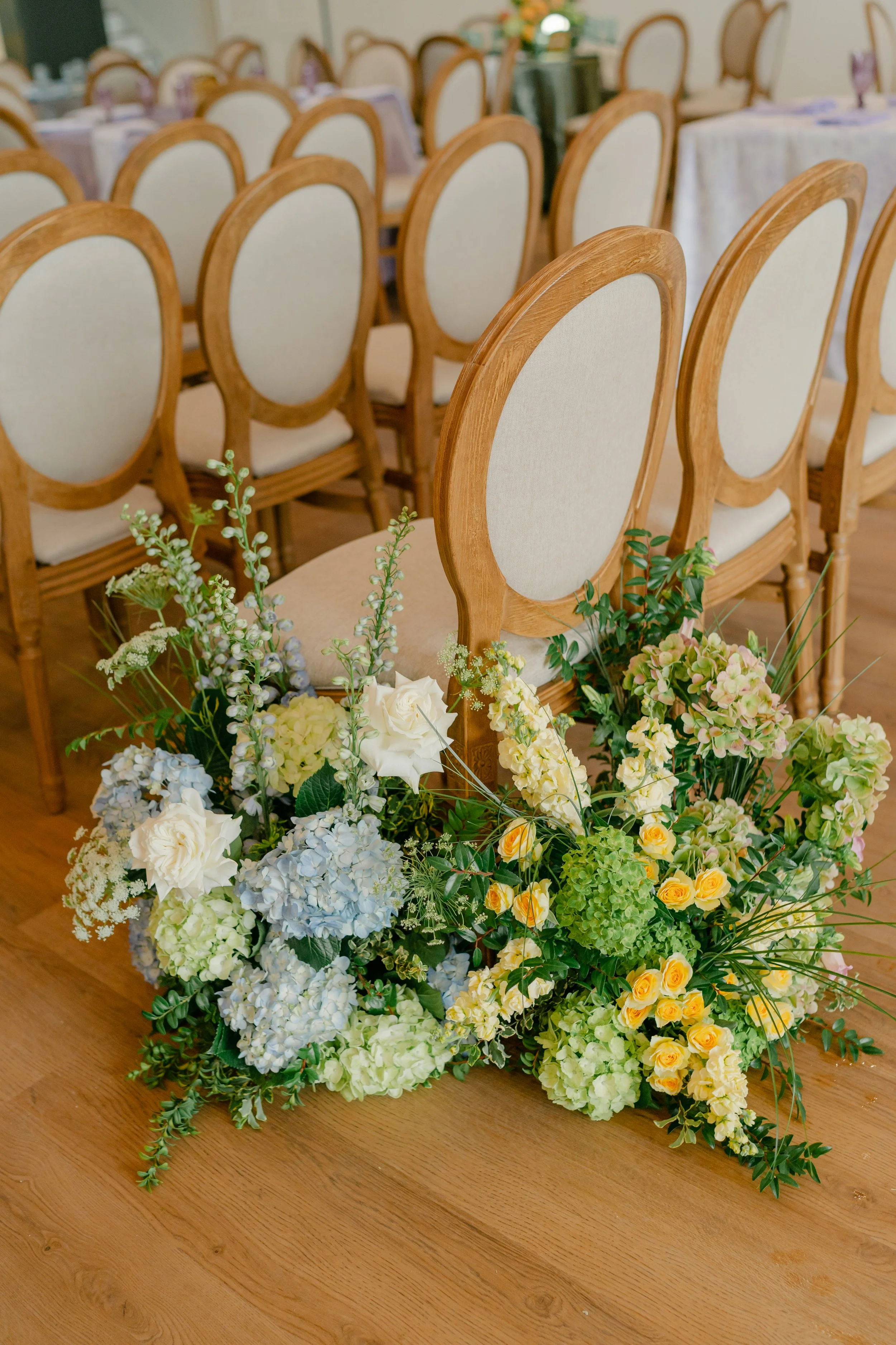 A large wedding floral arrangement with white, blue, and yellow flowers placed on a wooden floor in front of empty dining chairs with light-colored cushions and wooden frames, in a decorated banquet hall or restaurant setting.