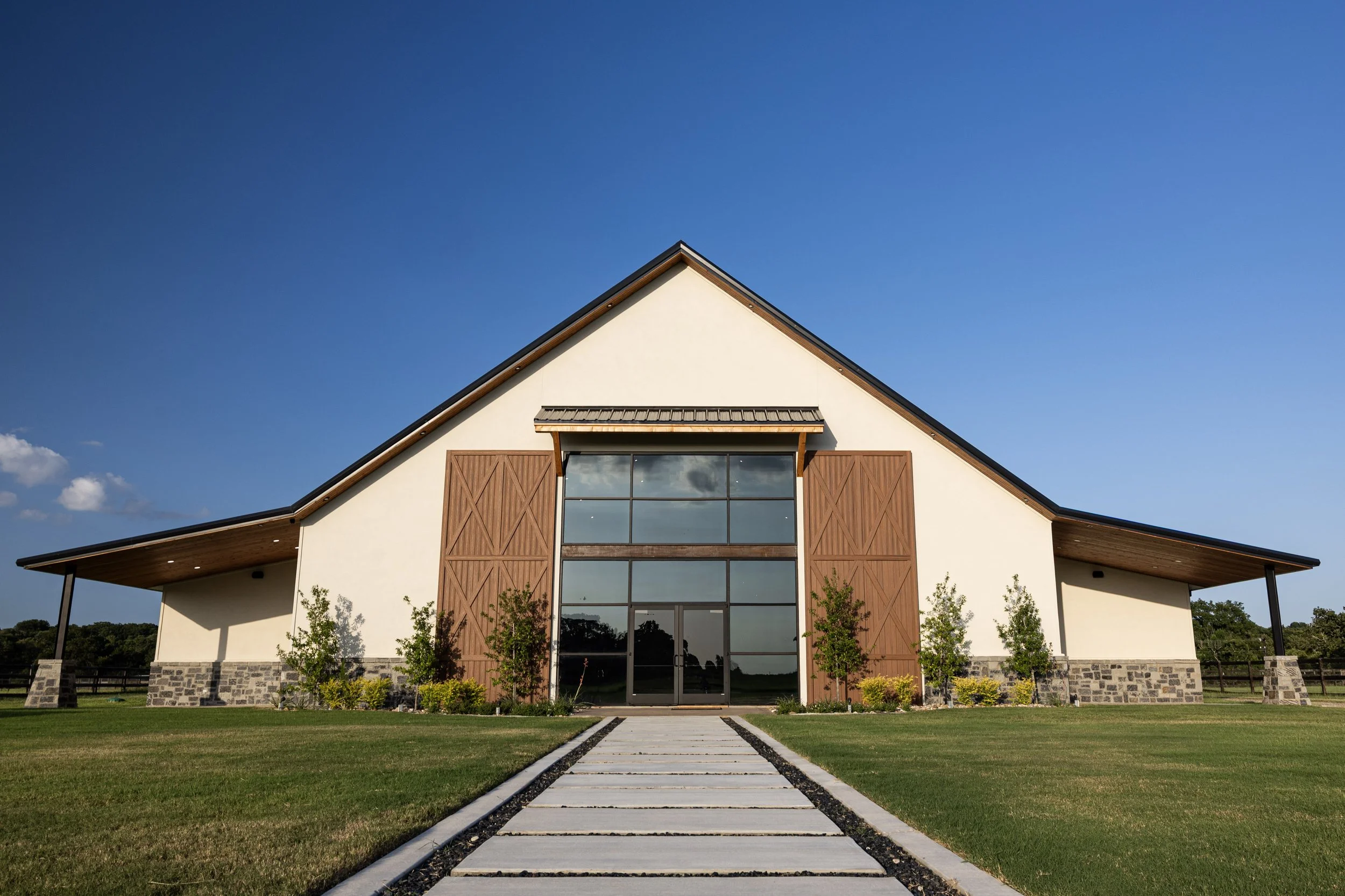 Modern house with a white facade, large glass windows, wooden accents, and a sharply peaked roof, surrounded by a green lawn and trees under a clear blue sky.