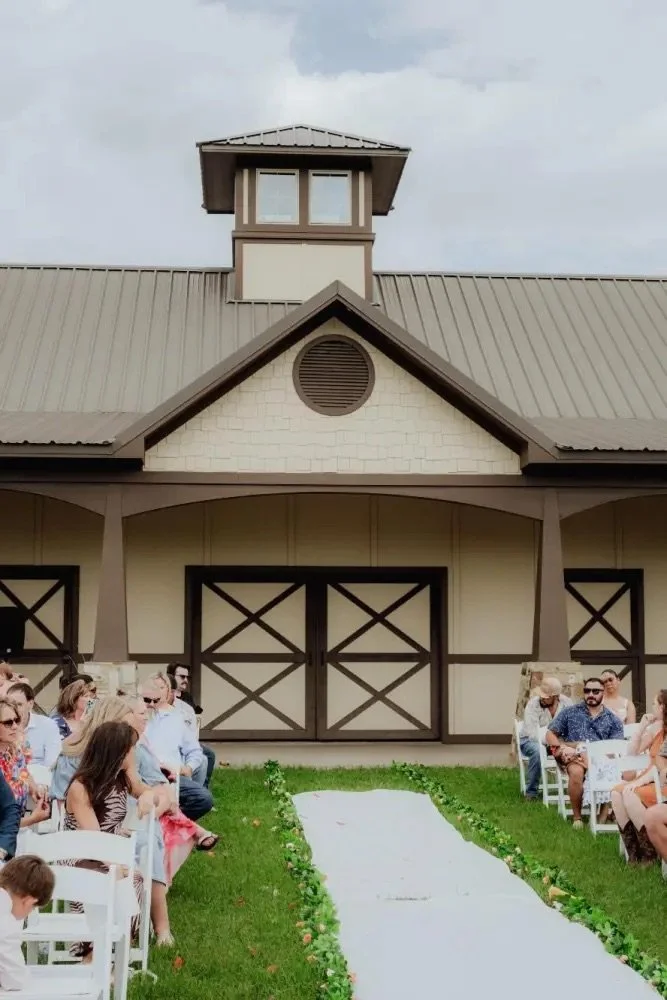 Outdoor wedding ceremony setup with people seated on both sides of a white aisle leading to an barn-style building with a large double door.