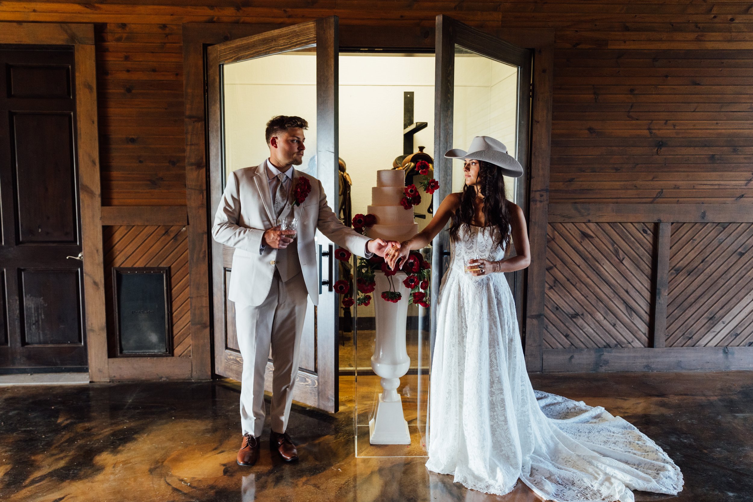 A couple in wedding attire holding hands in front of a decorative floral display during their wedding ceremony in a rustic indoor setting with wooden walls.