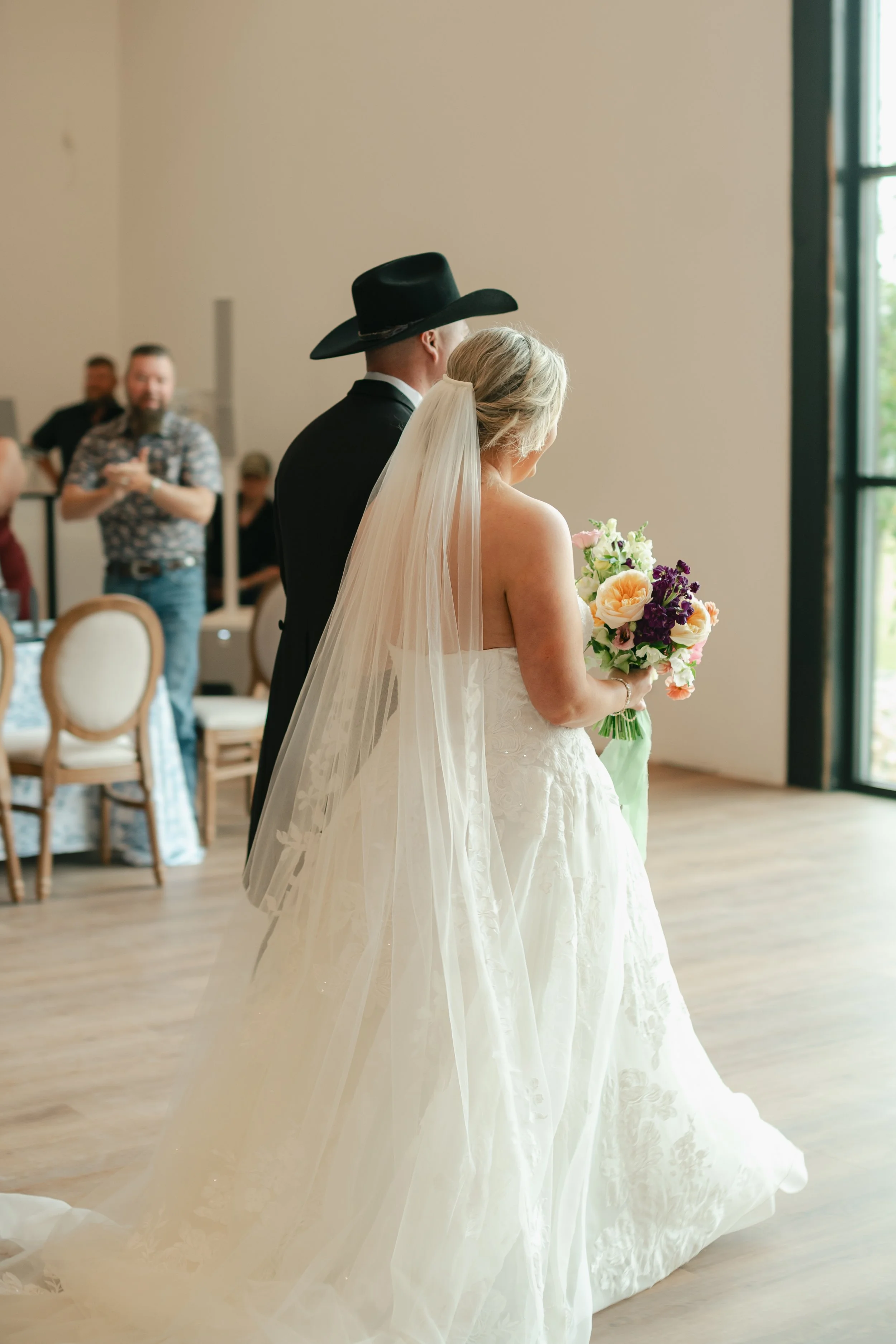 A bride and groom standing together during a wedding ceremony, with the bride holding a bouquet of flowers, inside a bright room with large windows and a wooden floor.