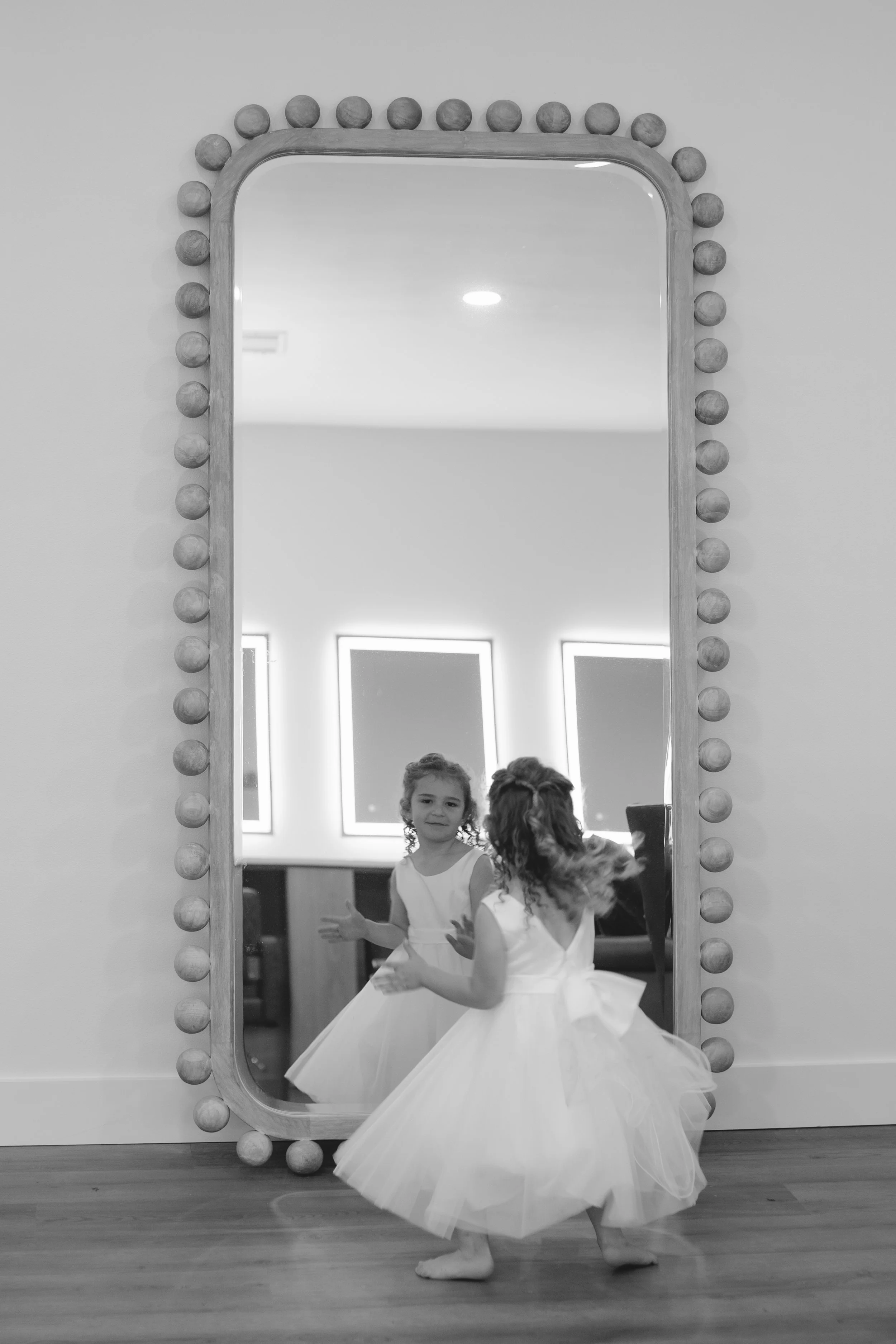 A young girl in a flower girl dress standing in front of a large mirror with a rounded frame decorated with round bulbs, inside a dance studio.