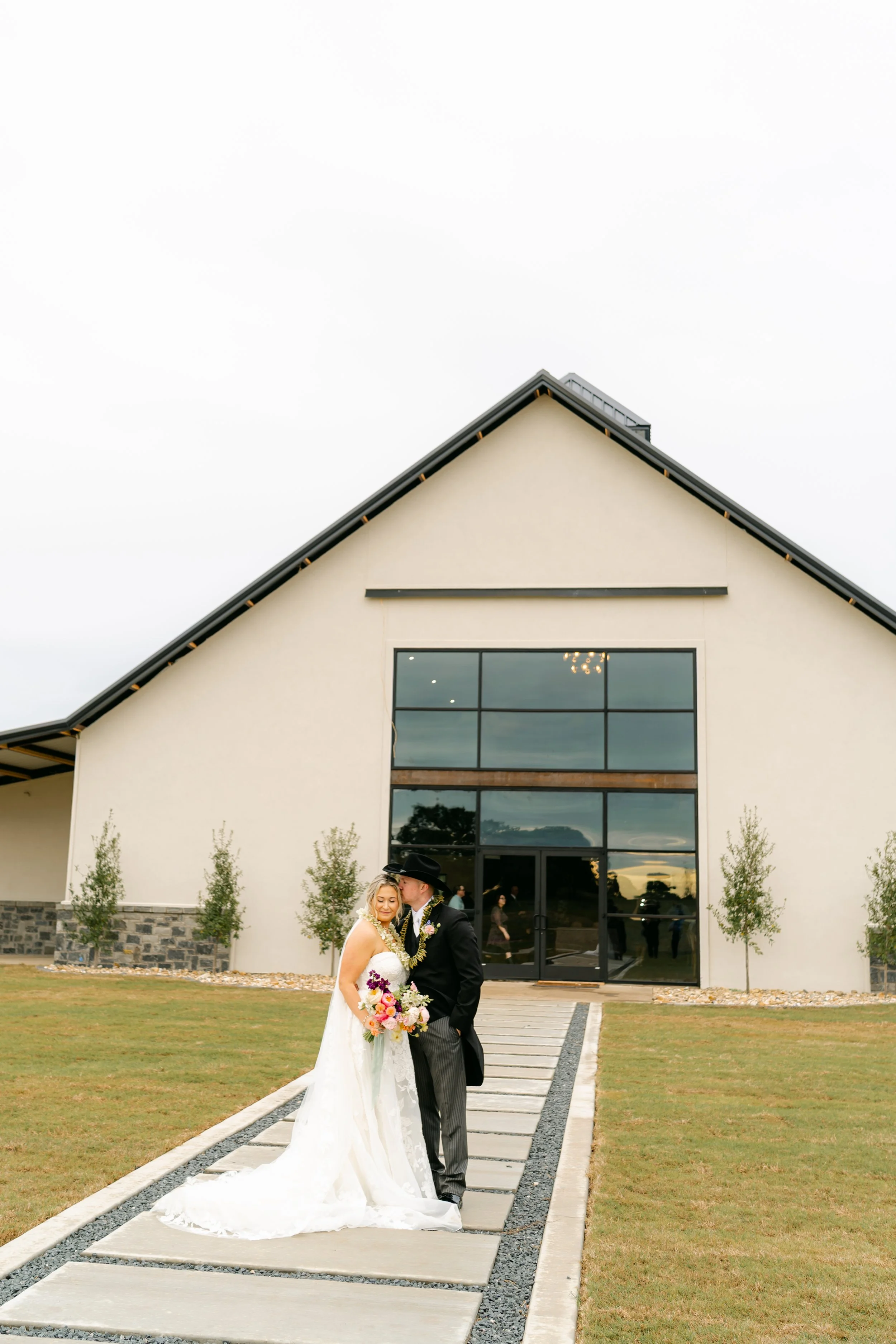 A newlywed couple standing on a walkway in front of a modern building with large glass windows, during a cloudy day. The bride is holding a colorful bouquet, and the groom is wearing a black cowboy hat.