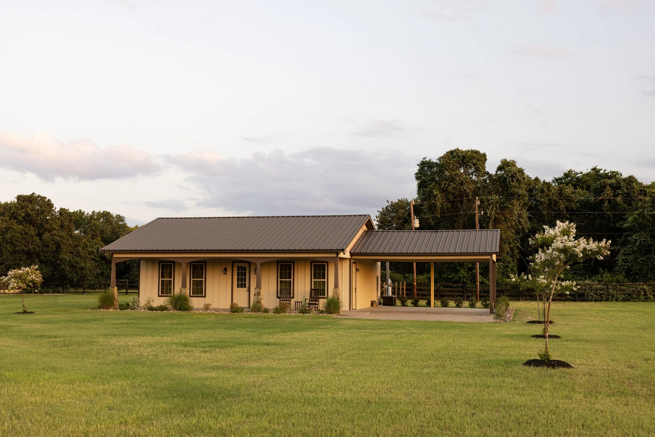 A yellow house with a metal roof, front porch with chairs, surrounded by green lawn and small trees, with a row of trees and a cloudy sky in the background.