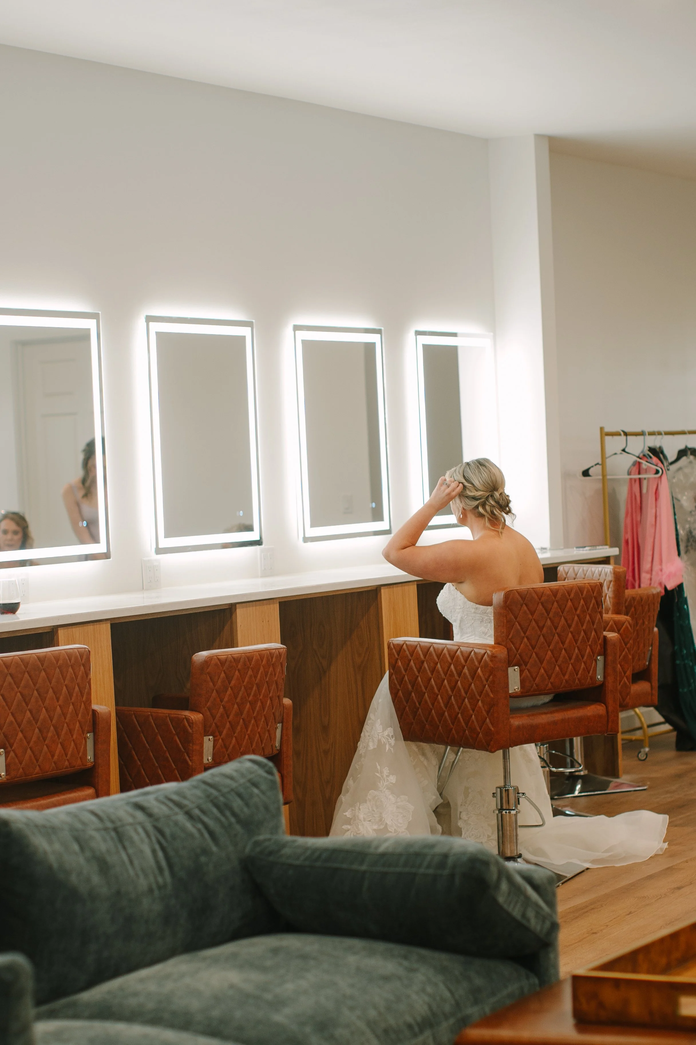 A bride seated in a salon chair in a wedding dress, adjusting her hair, with a modern vanity mirror and warm lighting in the background.