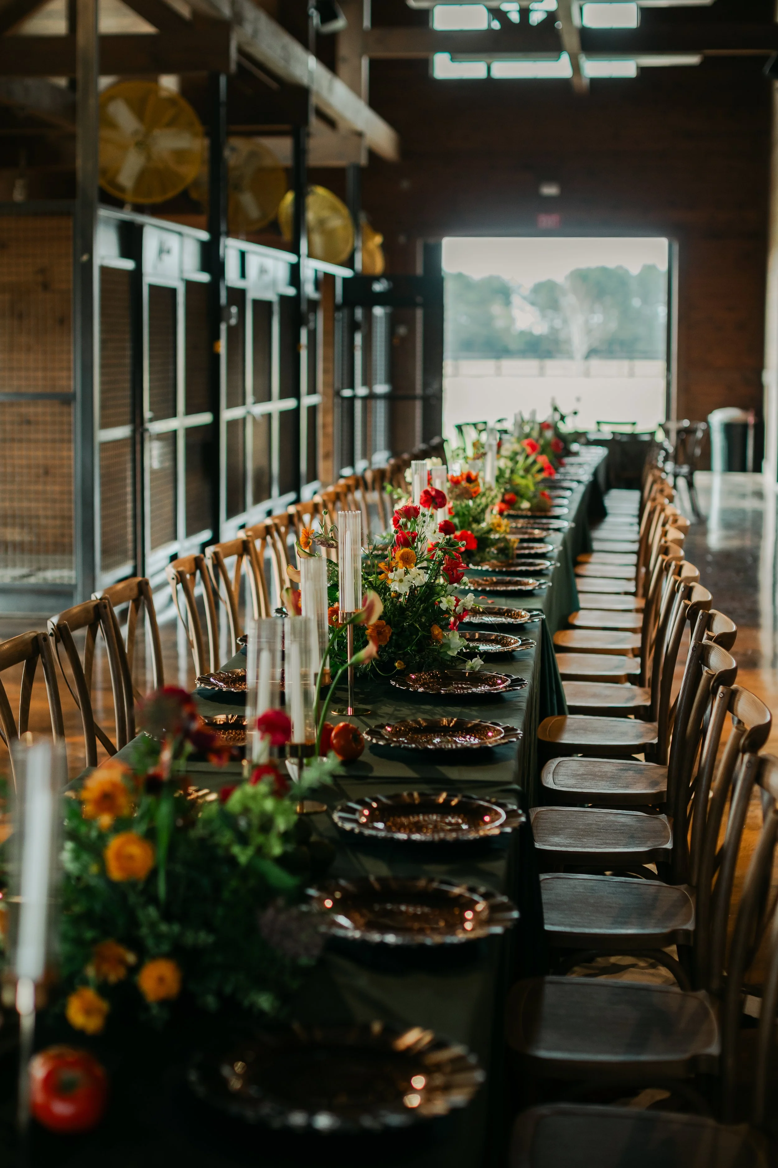 A long dining table set for an event, decorated with floral centerpieces and candleholders, inside a rustic wooden building with large windows and a view of trees outside.