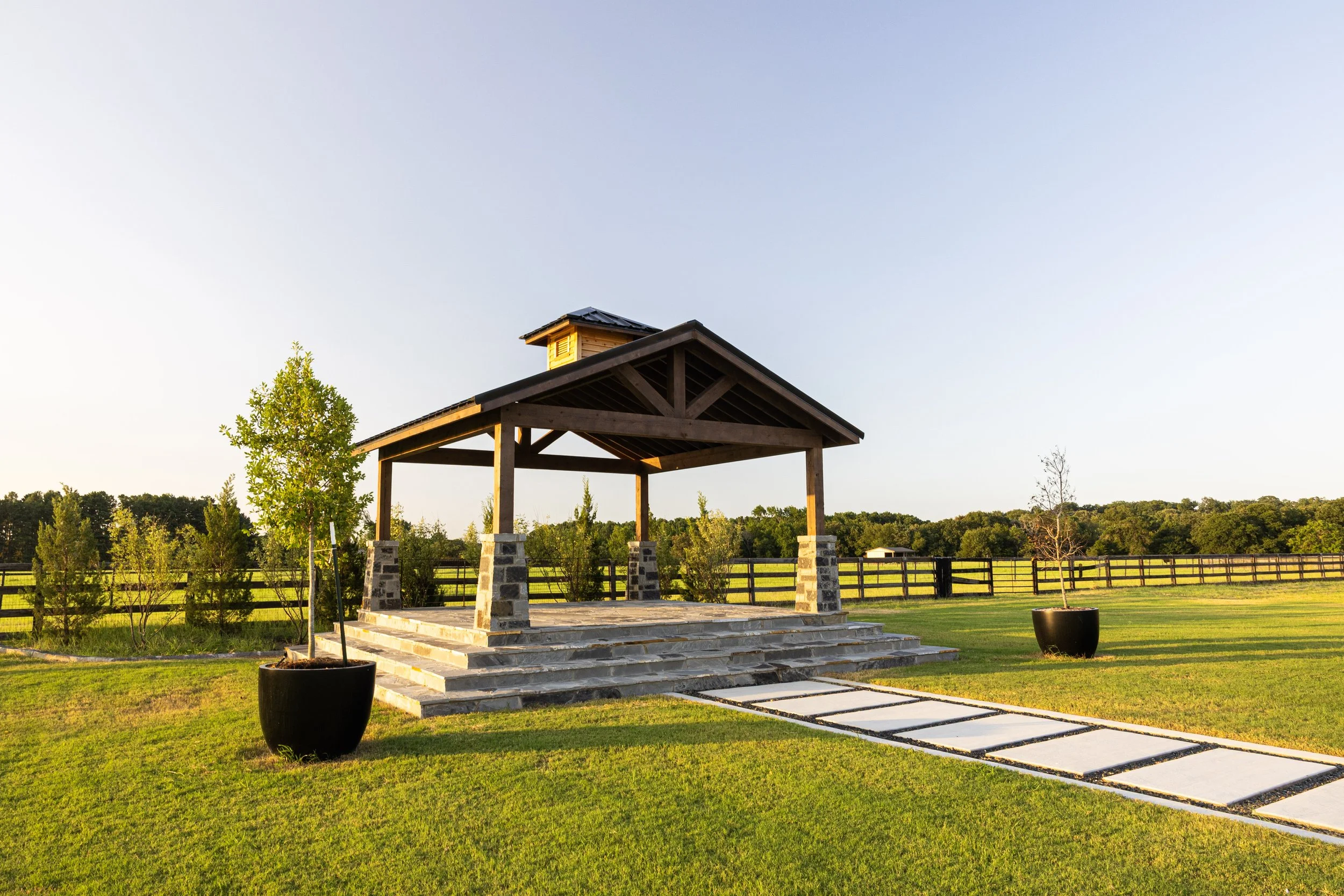 A wedding arbor gazebo with a shingled roof situated on a stone platform, surrounded by green grass, trees, and potted plants, under a clear blue sky.