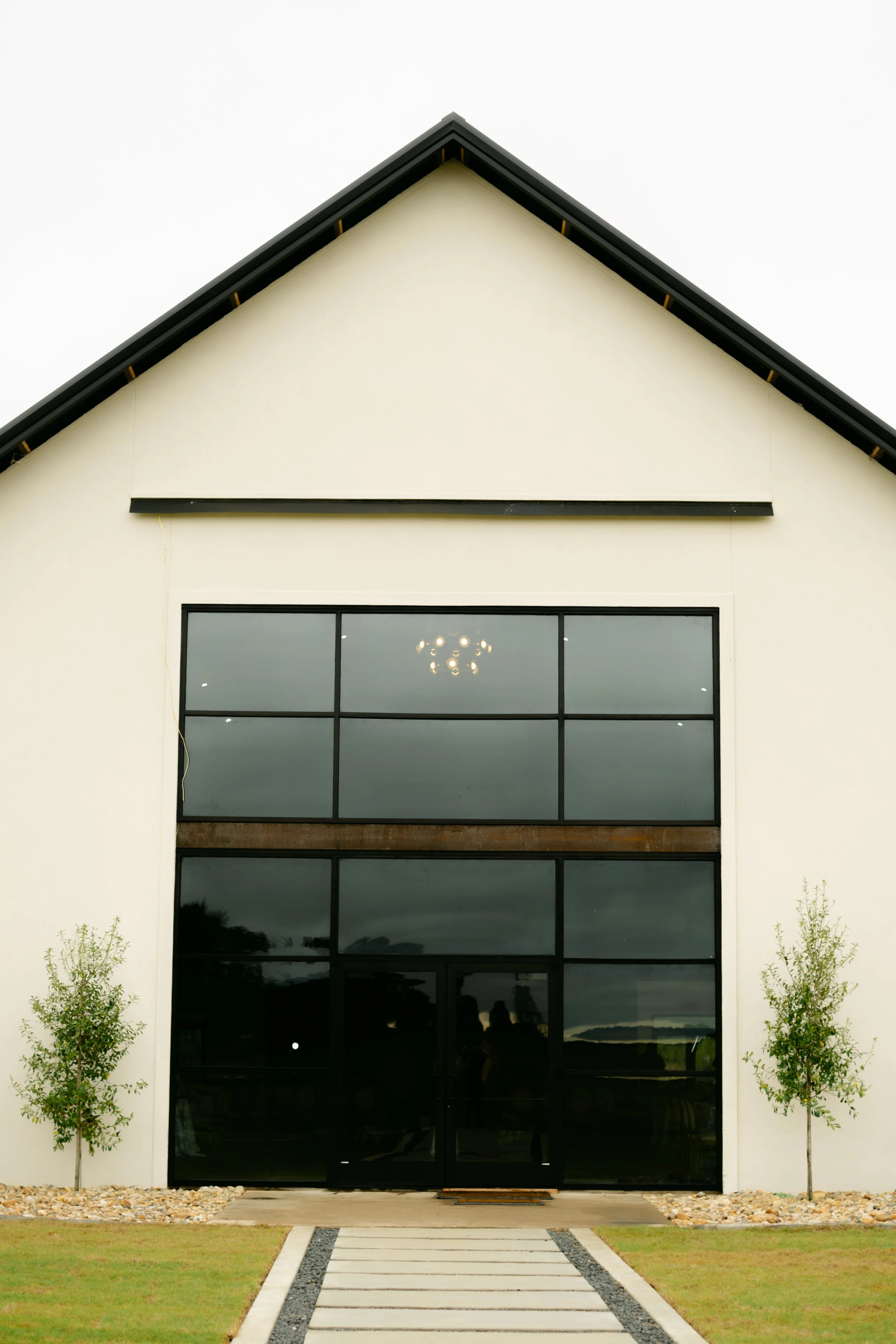 Front view of a modern building with a white facade, large black framed windows, and a pointed roof. Small trees are planted on either side of the walkway leading to the entrance on a cloudy day.