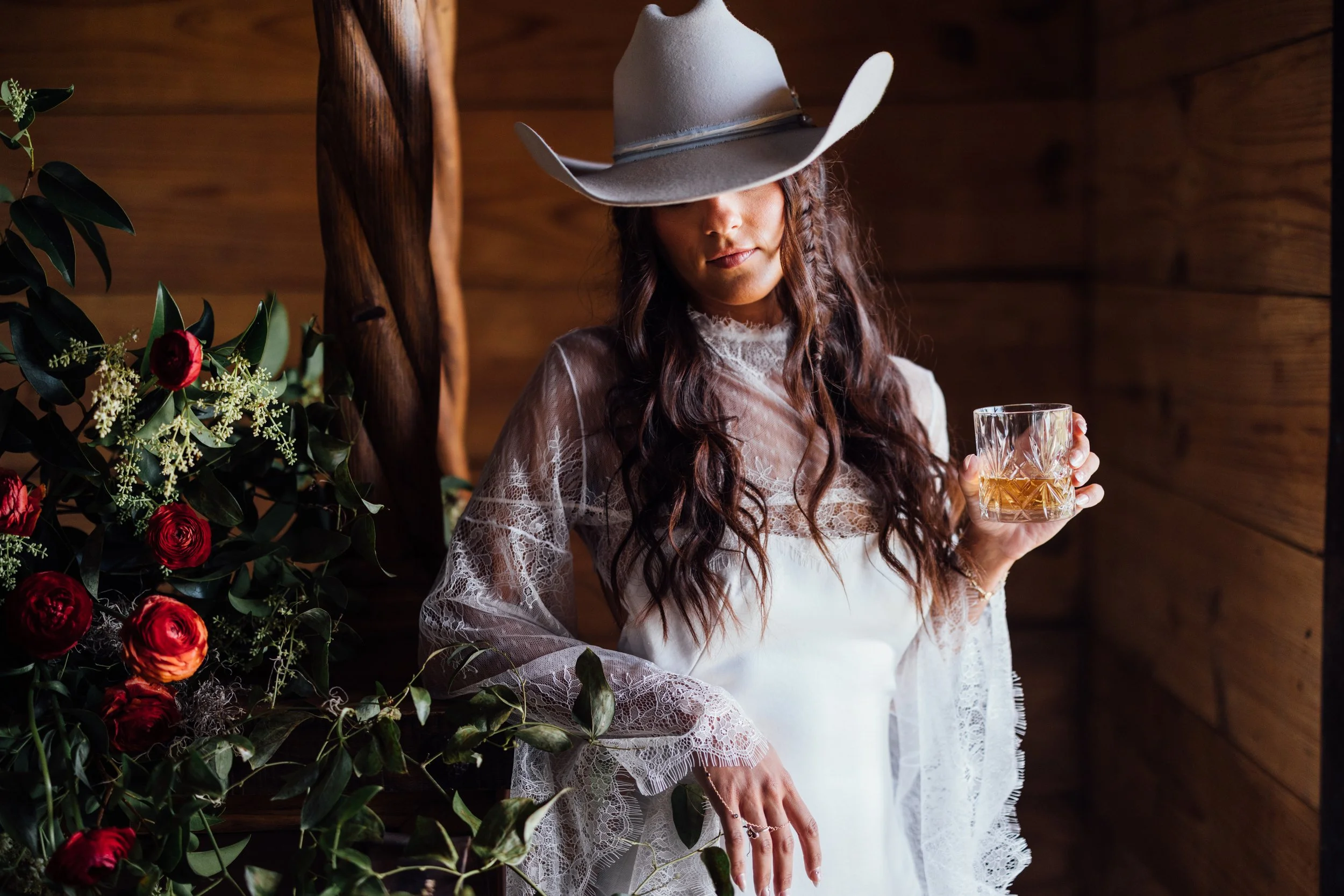 A woman wearing a white lace wedding dress and a wide-brimmed cowboy hat holding a glass of whiskey, standing beside a floral arrangement in a rustic wooden setting.