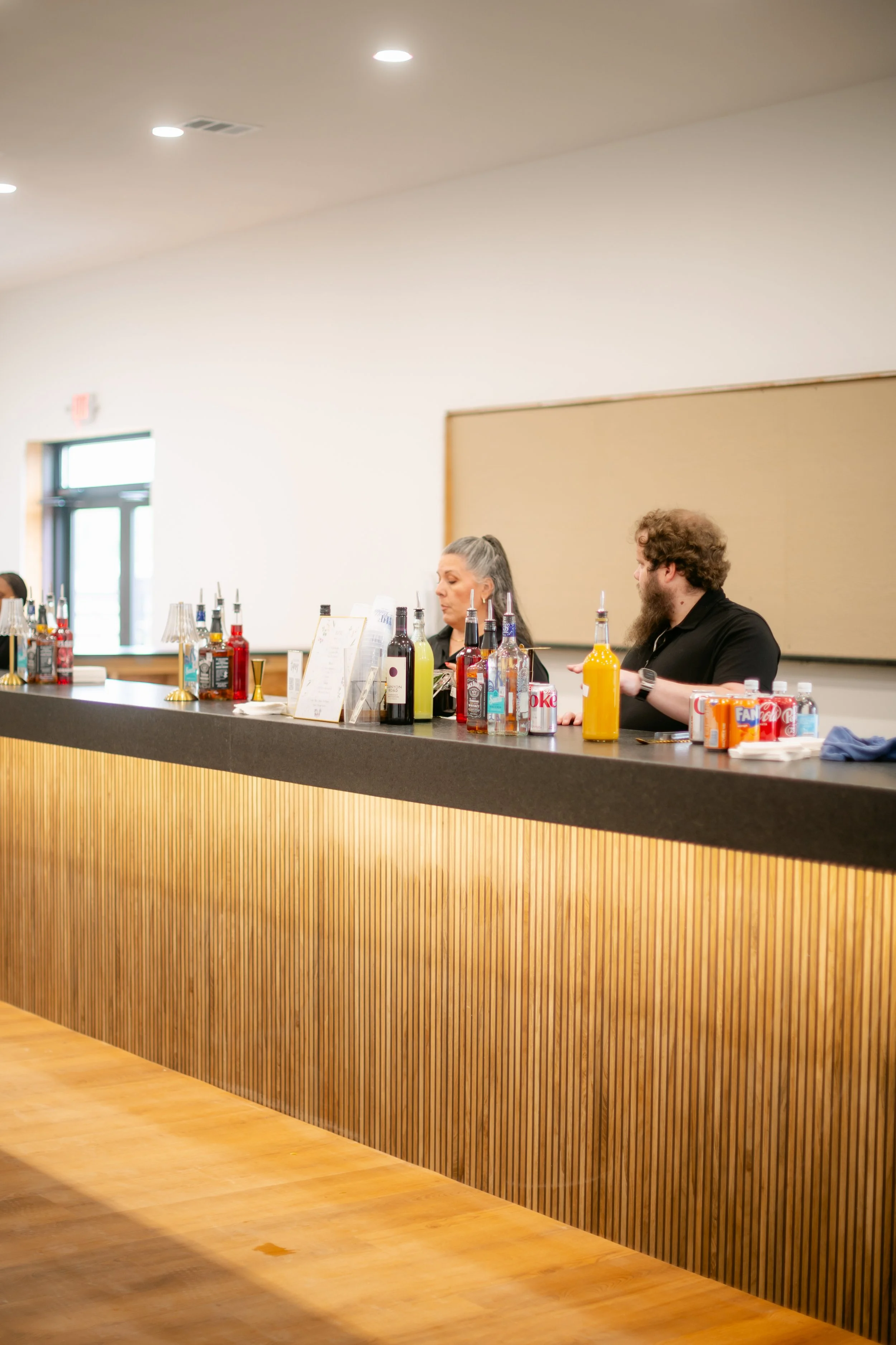Bar counter with various liquor bottles, soda cans, and glasses, with two people serving behind the counter in a bright, modern room.