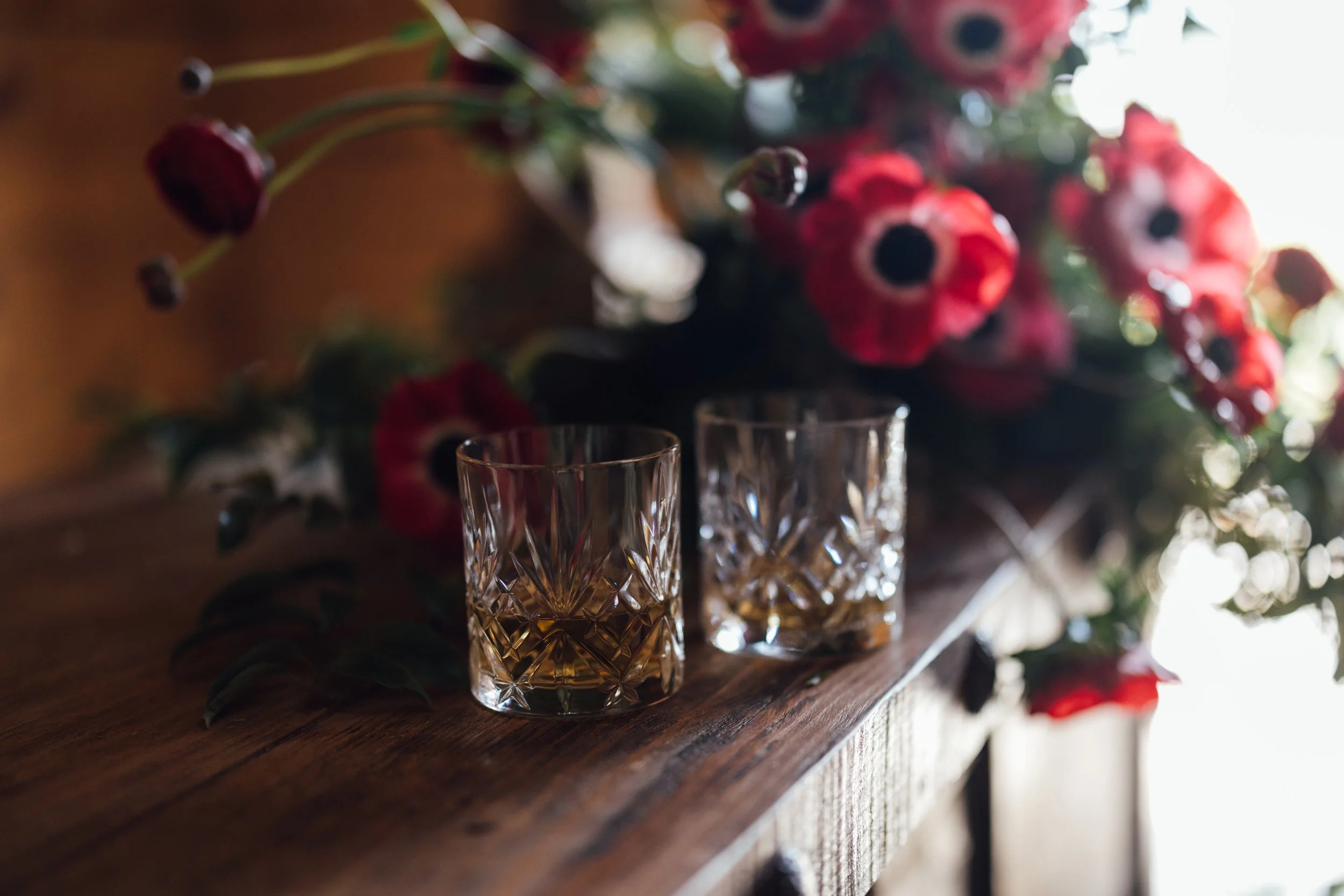 Close-up of two cut glass glasses with a diamond pattern on a wooden surface, with a blurred background of red flowers and greenery.