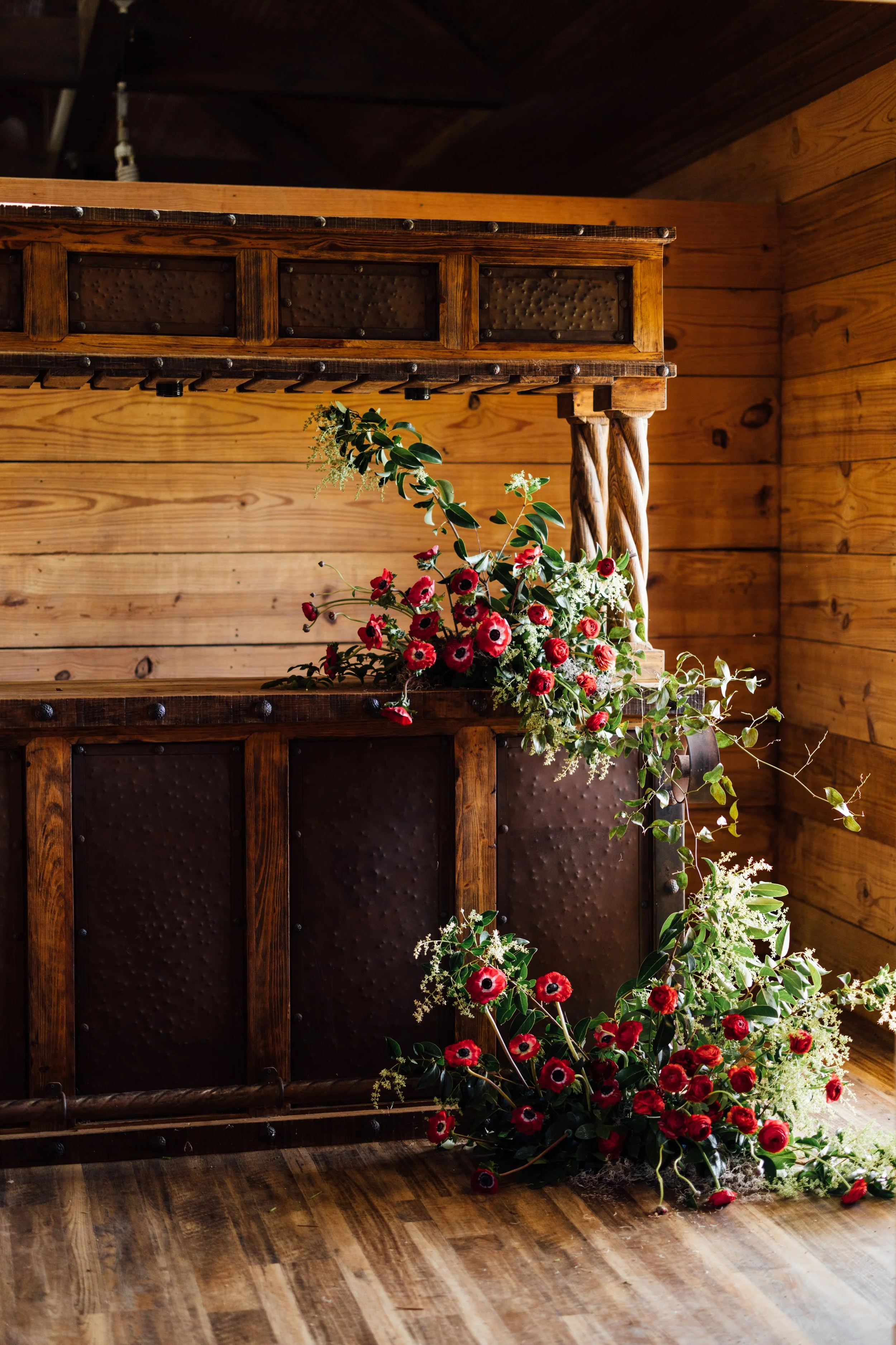 A wooden mantle with black metal accents decorated with a bouquet of red and white flowers and greenery against a wooden wall.
