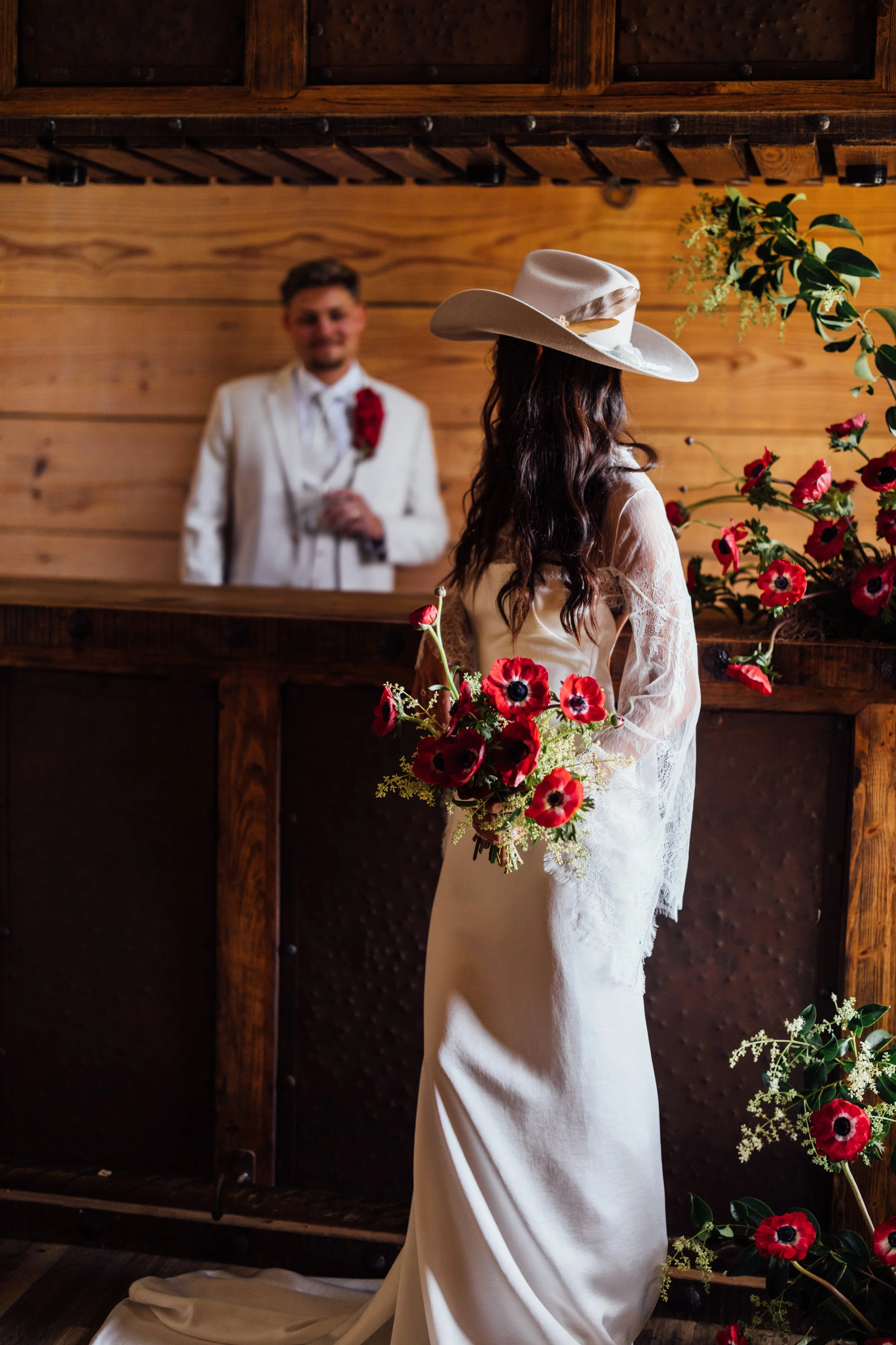 A woman dressed in a white lace wedding gown and wearing a wide-brimmed hat is holding a bouquet of vibrant red poppies with black centers, standing in front of a man in a white tuxedo with a red flower boutonniere, inside a wooden venue decorated wi