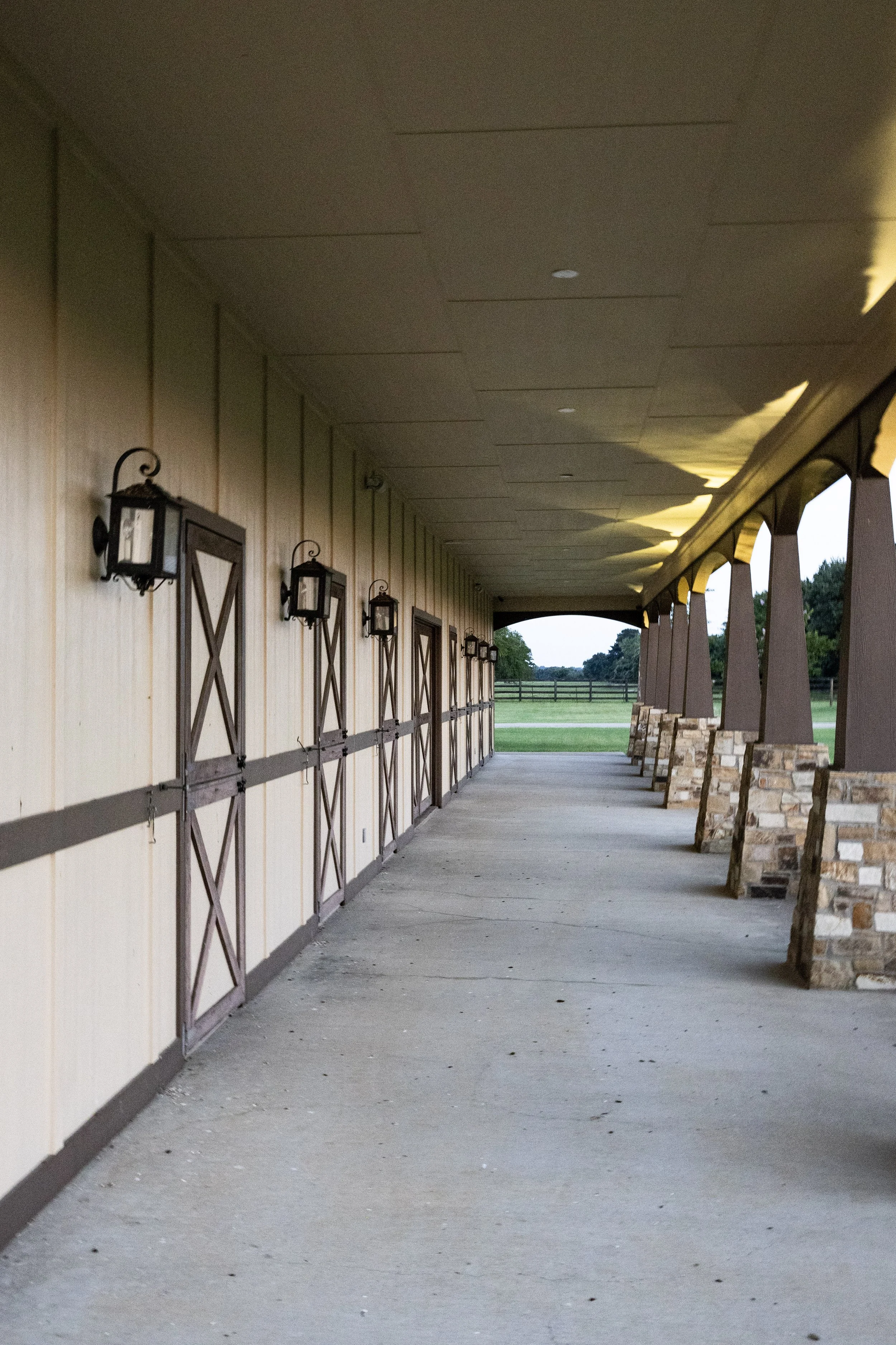Empty porch area with wall-mounted lanterns, barn-style doors, and a covered walkway leading to a rural field with trees in the distance.