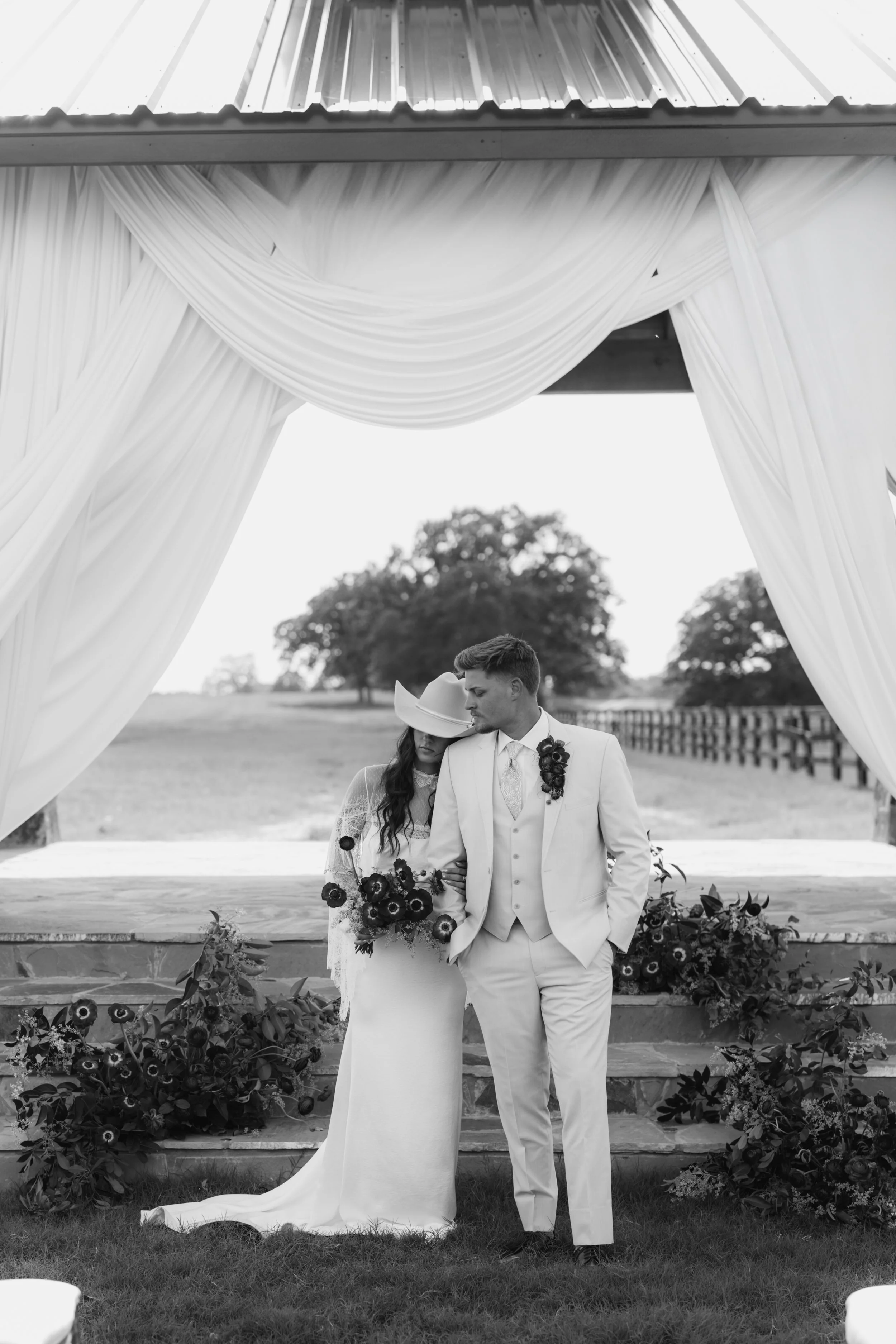 A black and white photo of a bride and groom standing together outside, under a decorated arch with draped fabric. The bride has long dark hair and is wearing a white wedding dress and hat, holding a bouquet. The groom is wearing a white suit and tie, with a boutonniere on his lapel, and has his hands in his pockets.