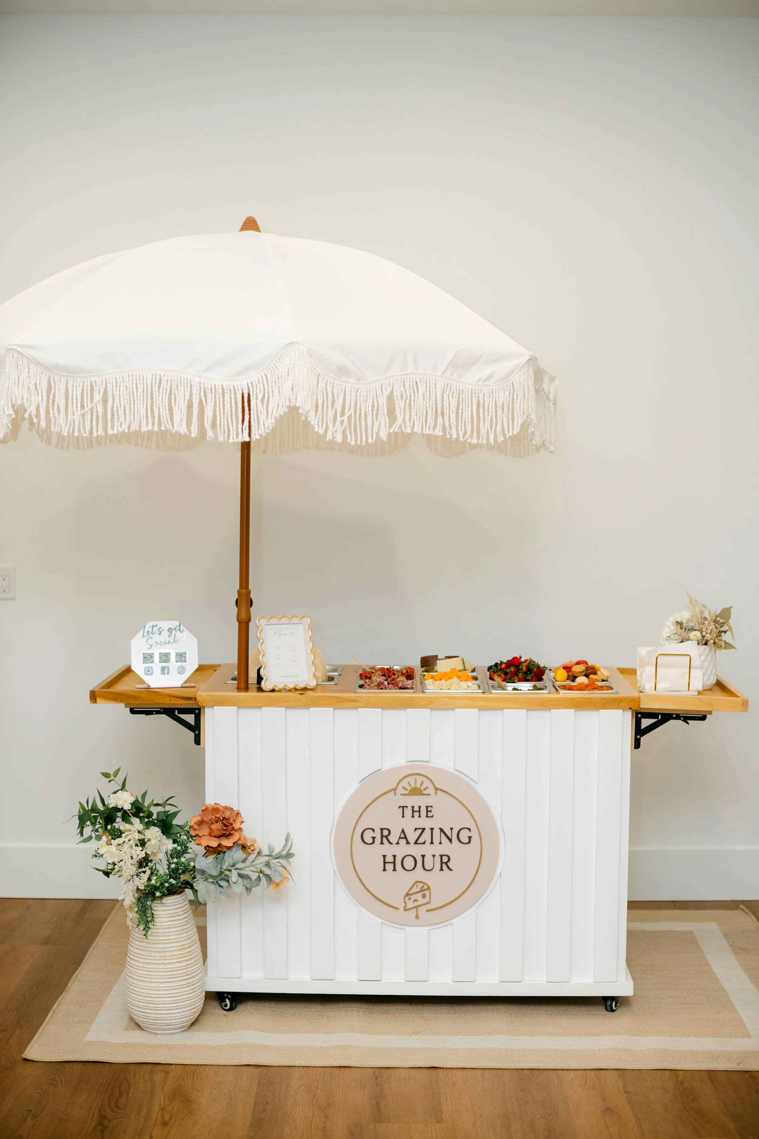 A white beverage umbrella with ruffled edges and a wooden pole, providing shade over a buffet table filled with cheese, strawberries, grapes, and other small foods, set against a plain wall. The table has a sign reading 'The Grazing Hour' and is deco