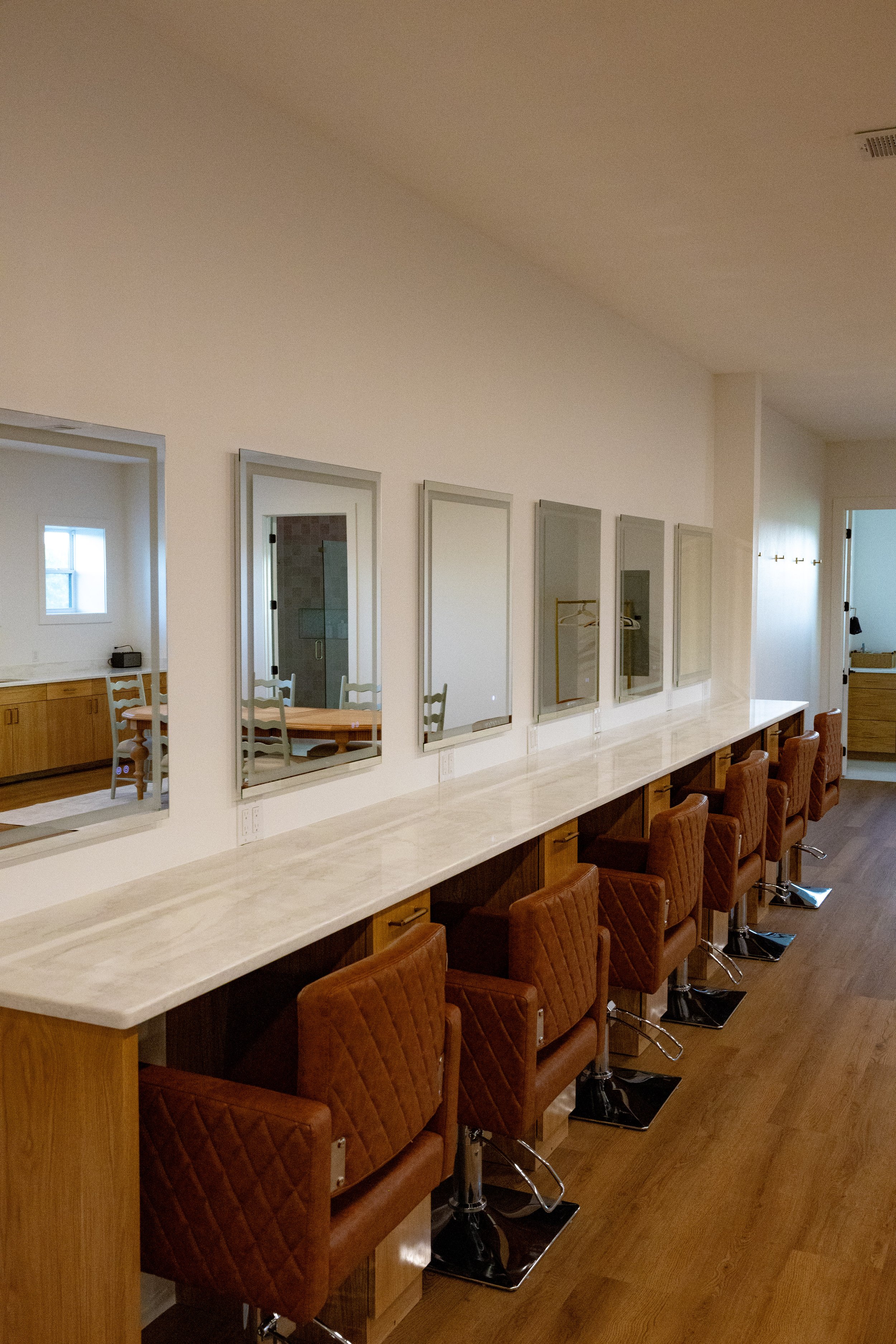 A row of brown leather salon chairs with quilted stitching along a long white counter with four mirrors on the wall behind it. The background shows a partial view of a dining area with a dining table and chairs through a doorway.